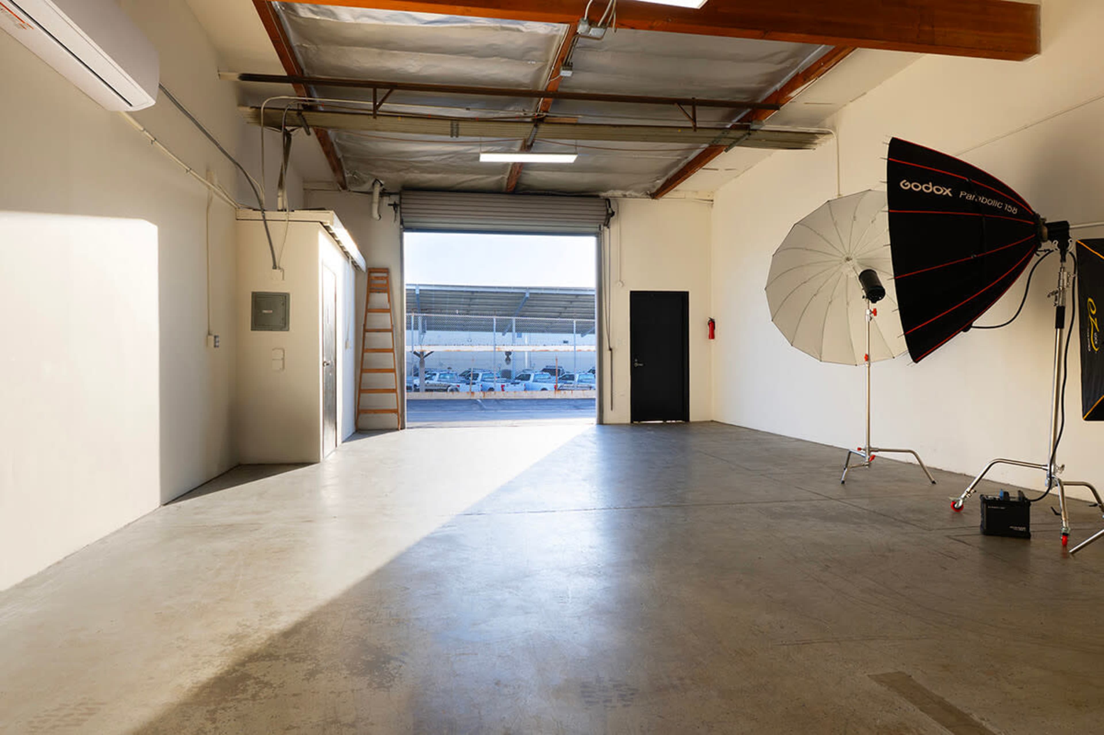 The image shows a spacious, empty studio interior with a large light umbrella and open access to an outdoor area beyond the doorway.