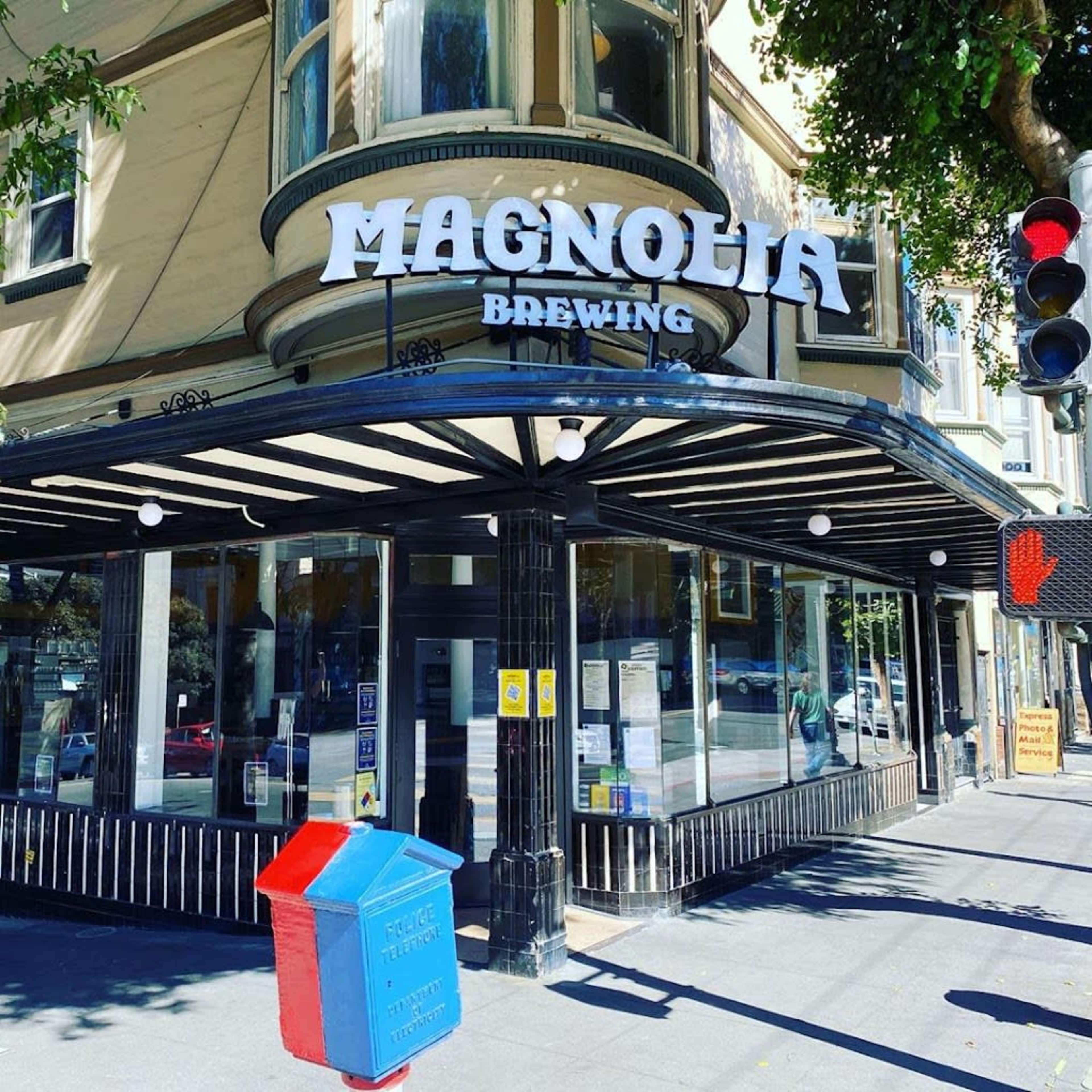 The image shows the exterior of Magnolia Brewing, featuring a large sign and a colorful trash bin at the corner of the building.