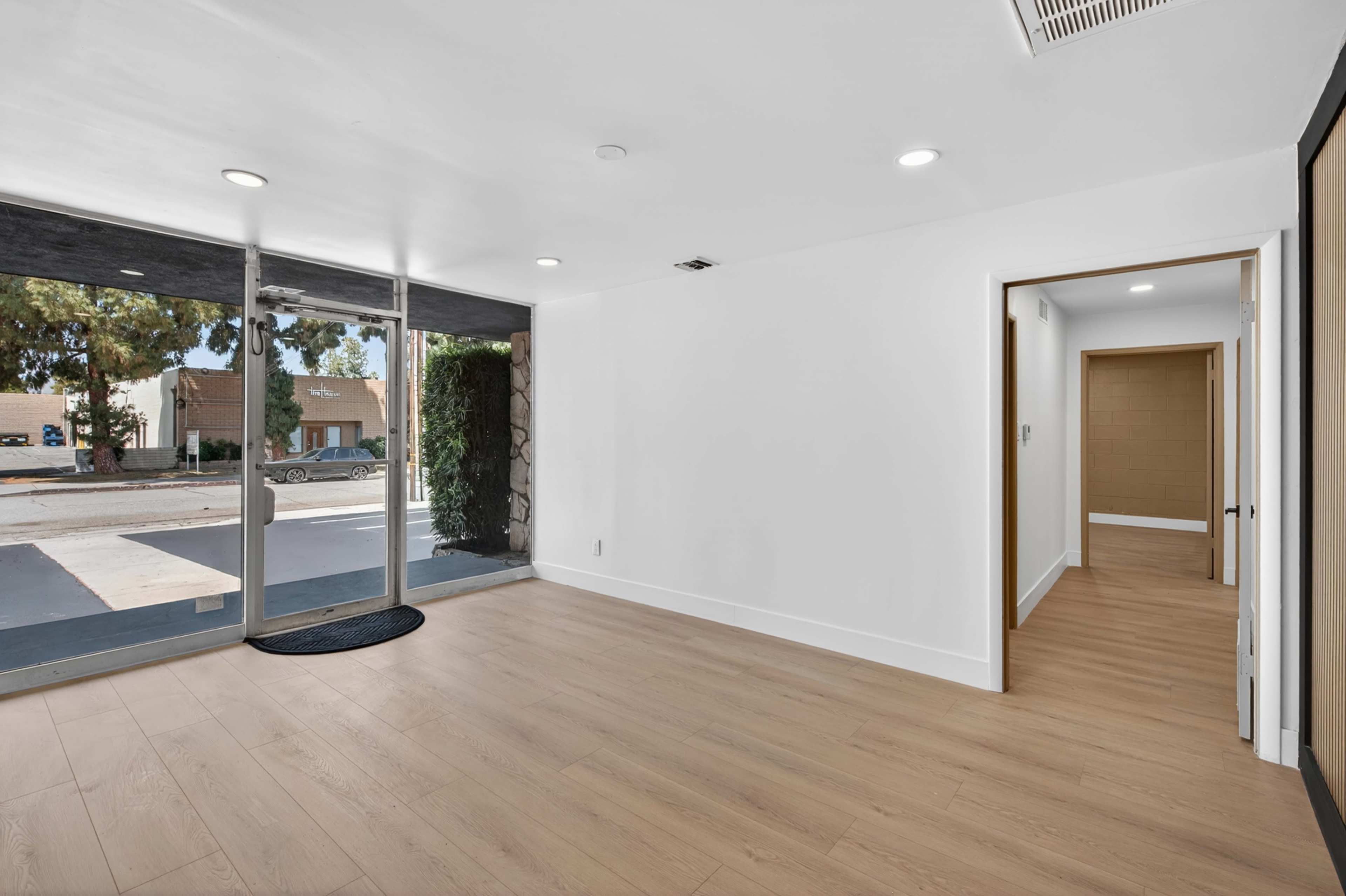 The image shows a modern, empty interior space with a large glass entrance and light wood flooring.