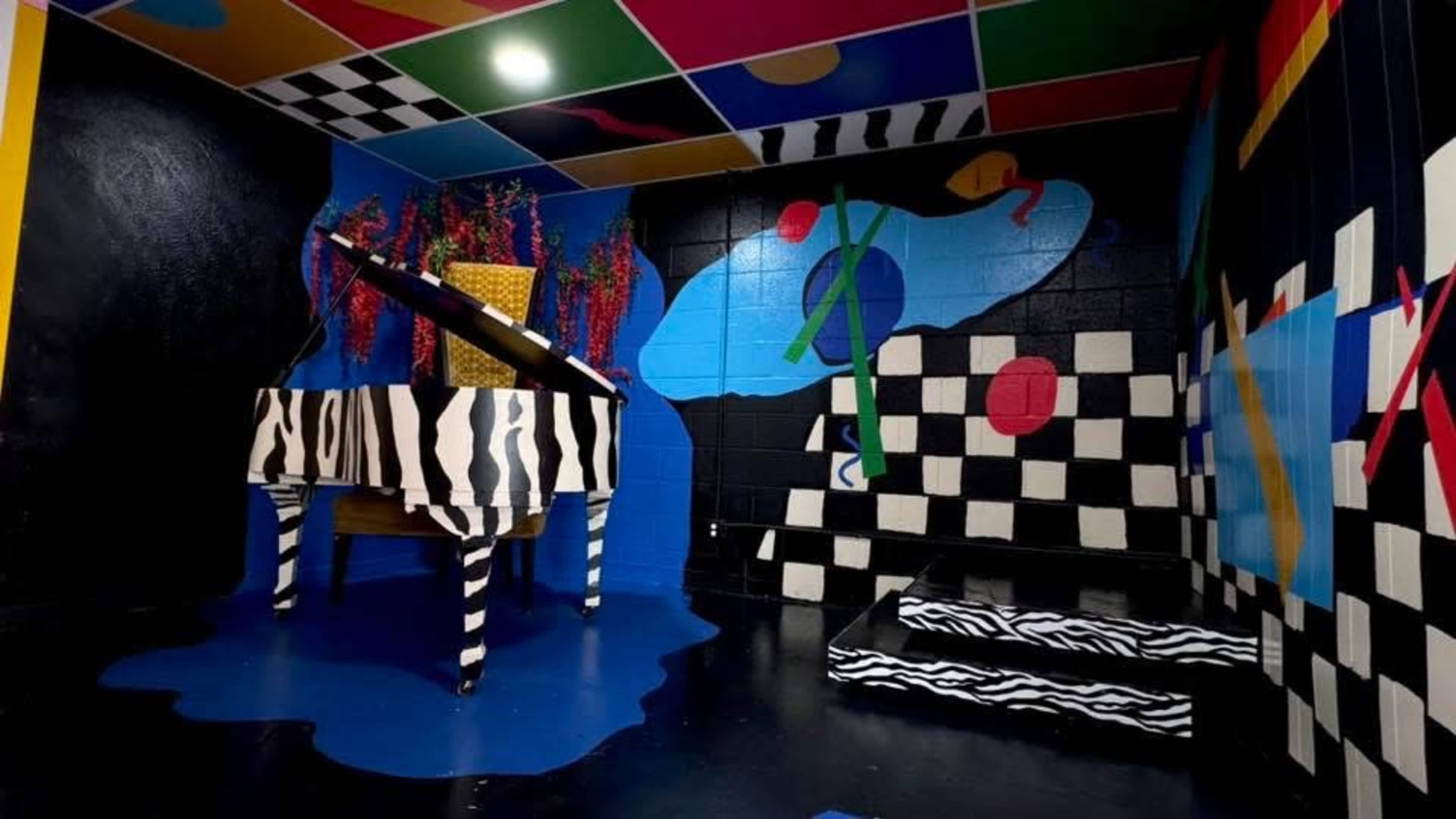 The image depicts a vibrant room with geometric patterns on the walls and floor, featuring a zebra-striped piano and colorful decorations.