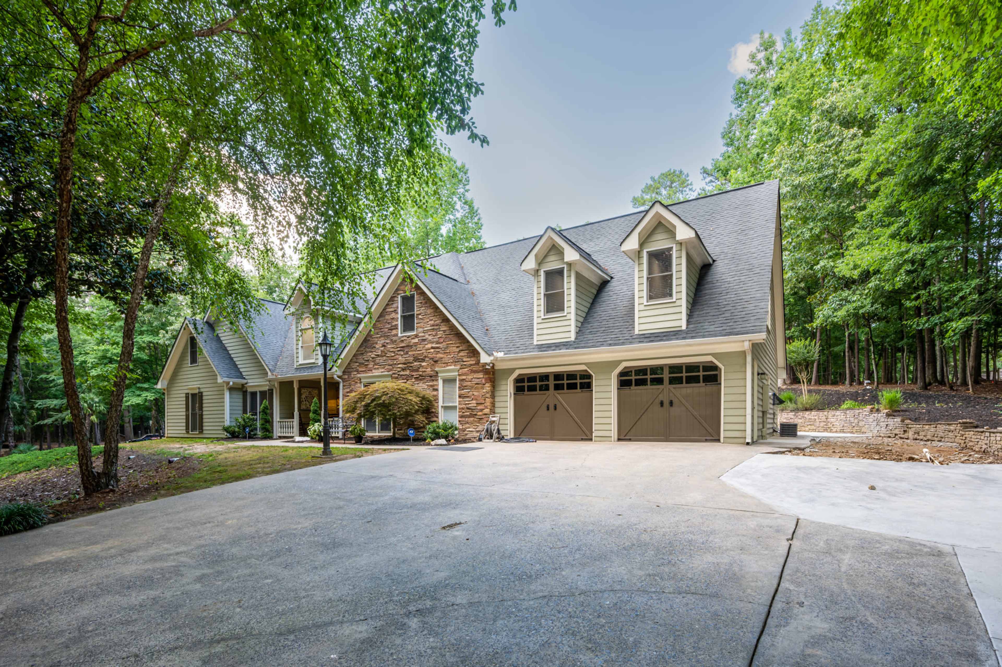 A large, two-story house with a stone and wooden facade is situated under trees, featuring a two-car garage and a concrete driveway.