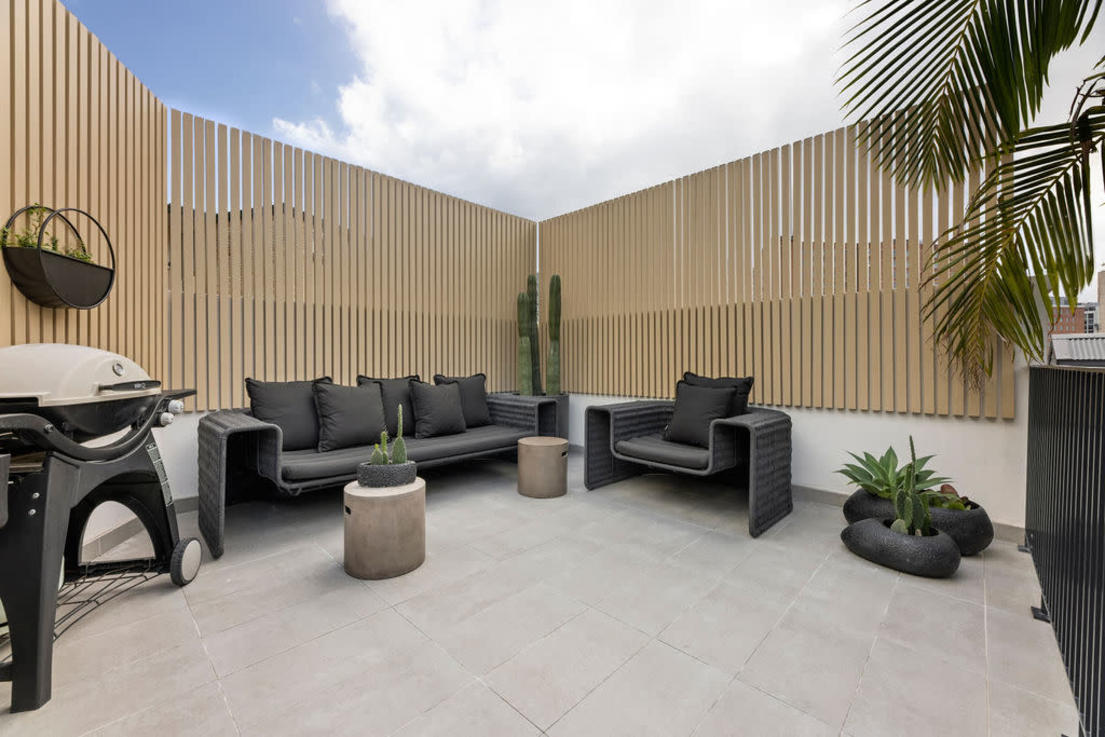 A modern outdoor terrace featuring a black sectional sofa, a small round table, a barbecue grill, and potted plants, all surrounded by wooden slat fencing.