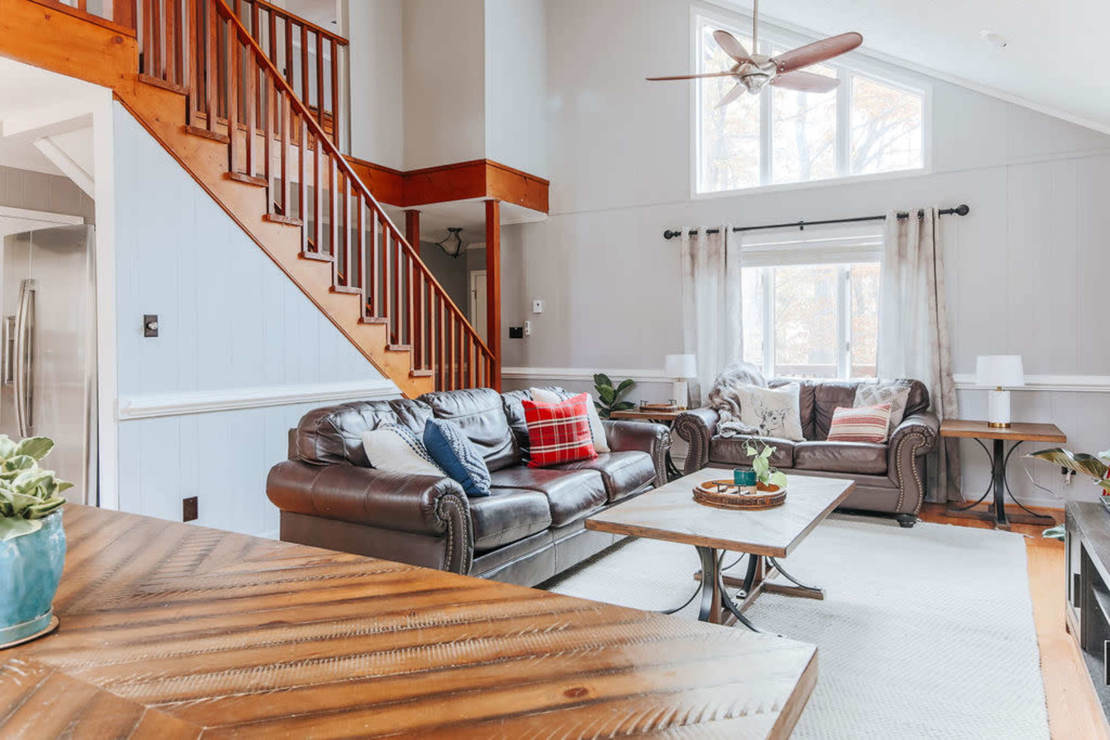 A cozy living room with a leather sofa and armchair, a wooden coffee table, and large windows allowing natural light to enter the space.