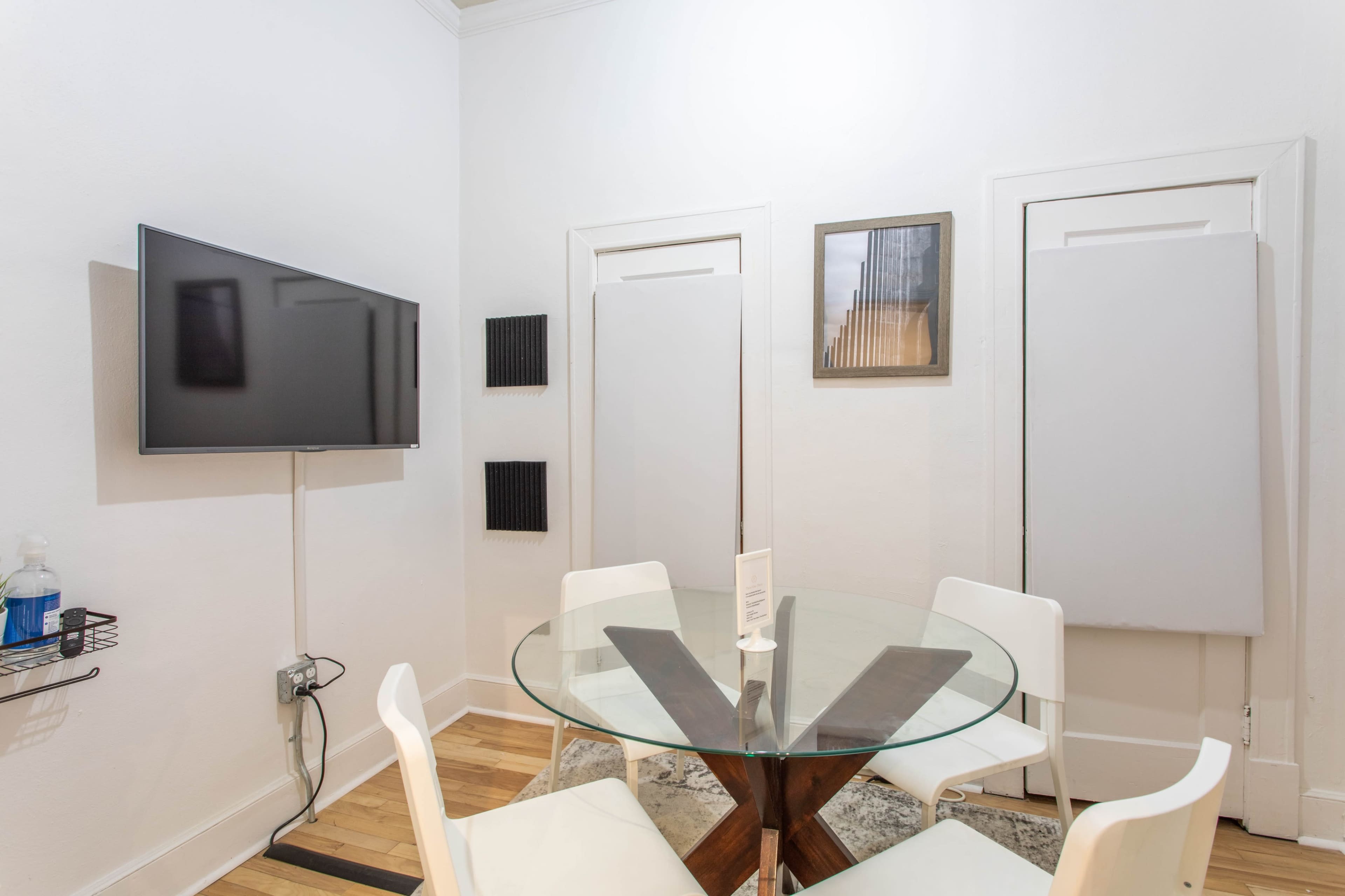 A round glass dining table with white chairs is set in a small room featuring a wall-mounted TV and two closed doors.