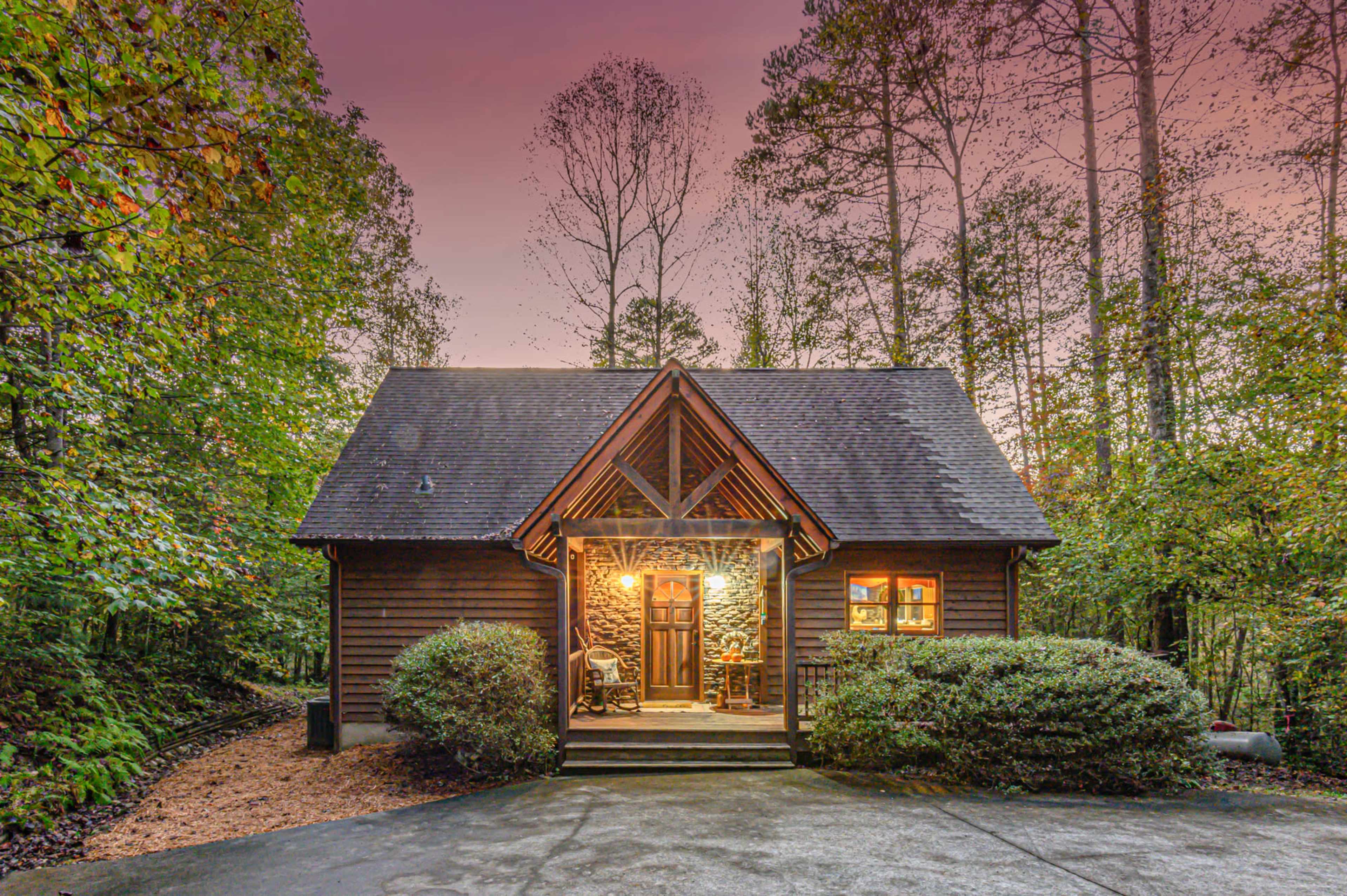 A wooden cabin with a sloped roof is situated amid trees and shrubs, featuring a welcoming front porch and a warmly lit entrance.