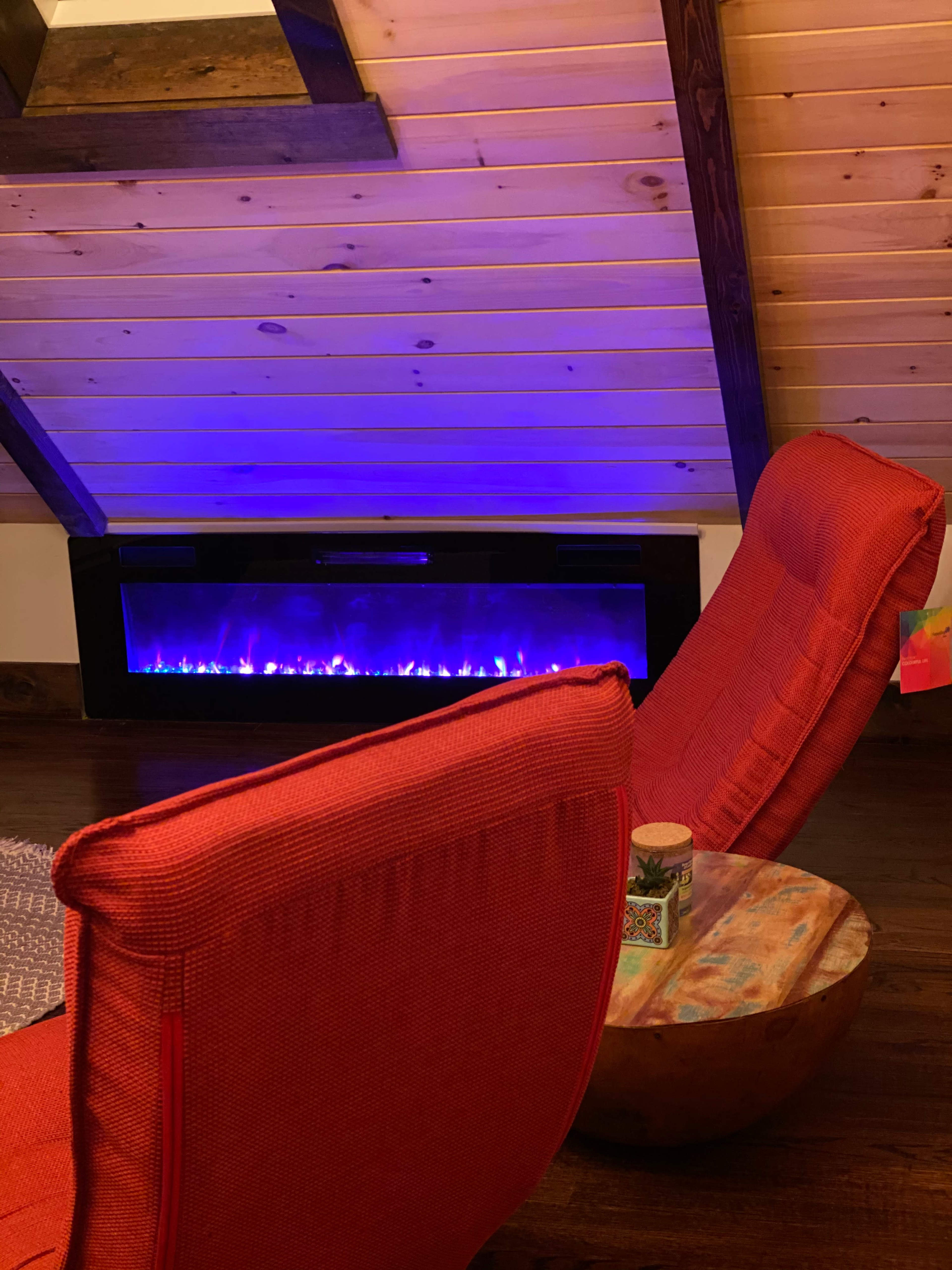 A cozy room features two red chairs facing an electric fireplace with blue and purple flames.