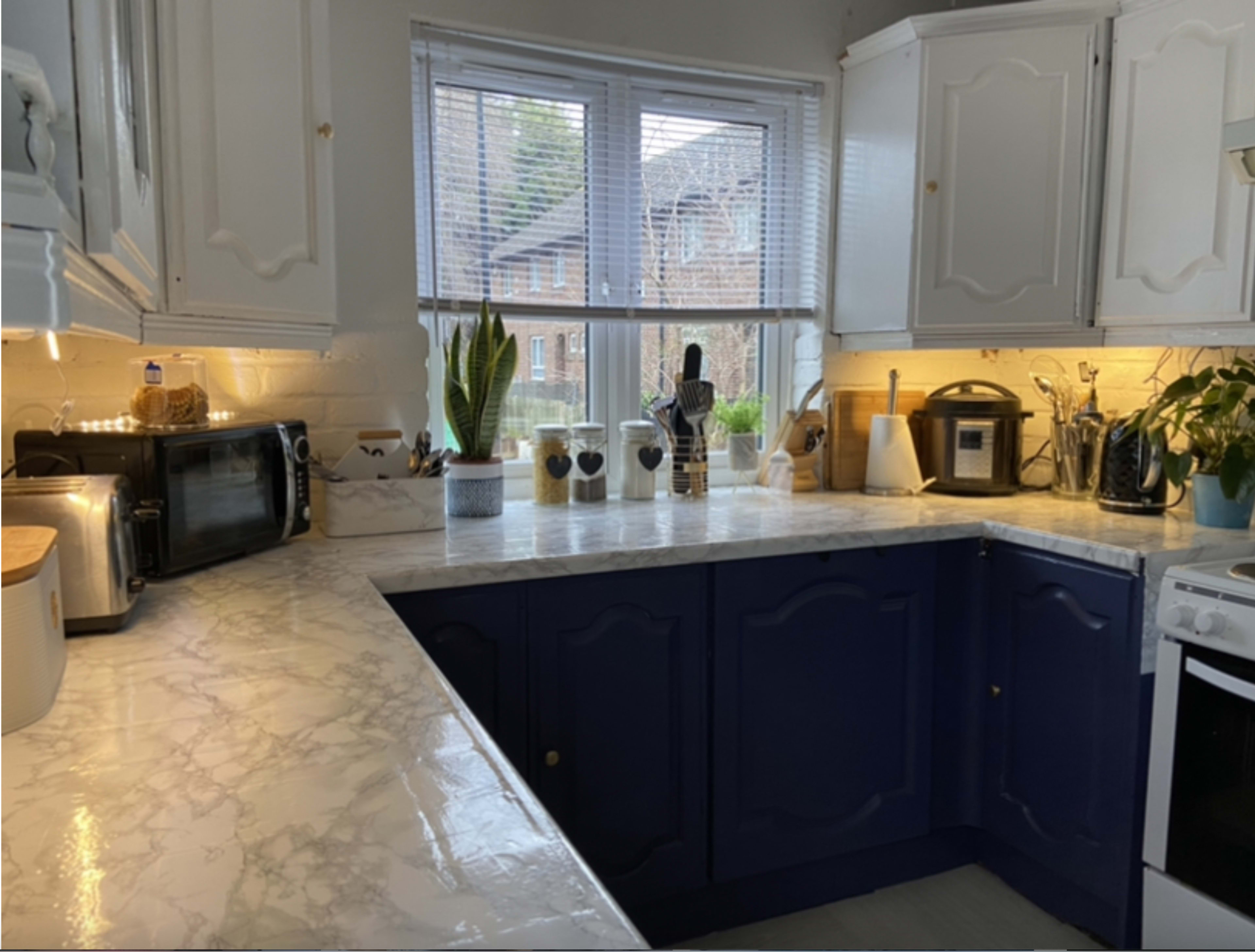 A kitchen with white and navy blue cabinetry, a marble countertop, and various kitchen appliances arranged around a window.