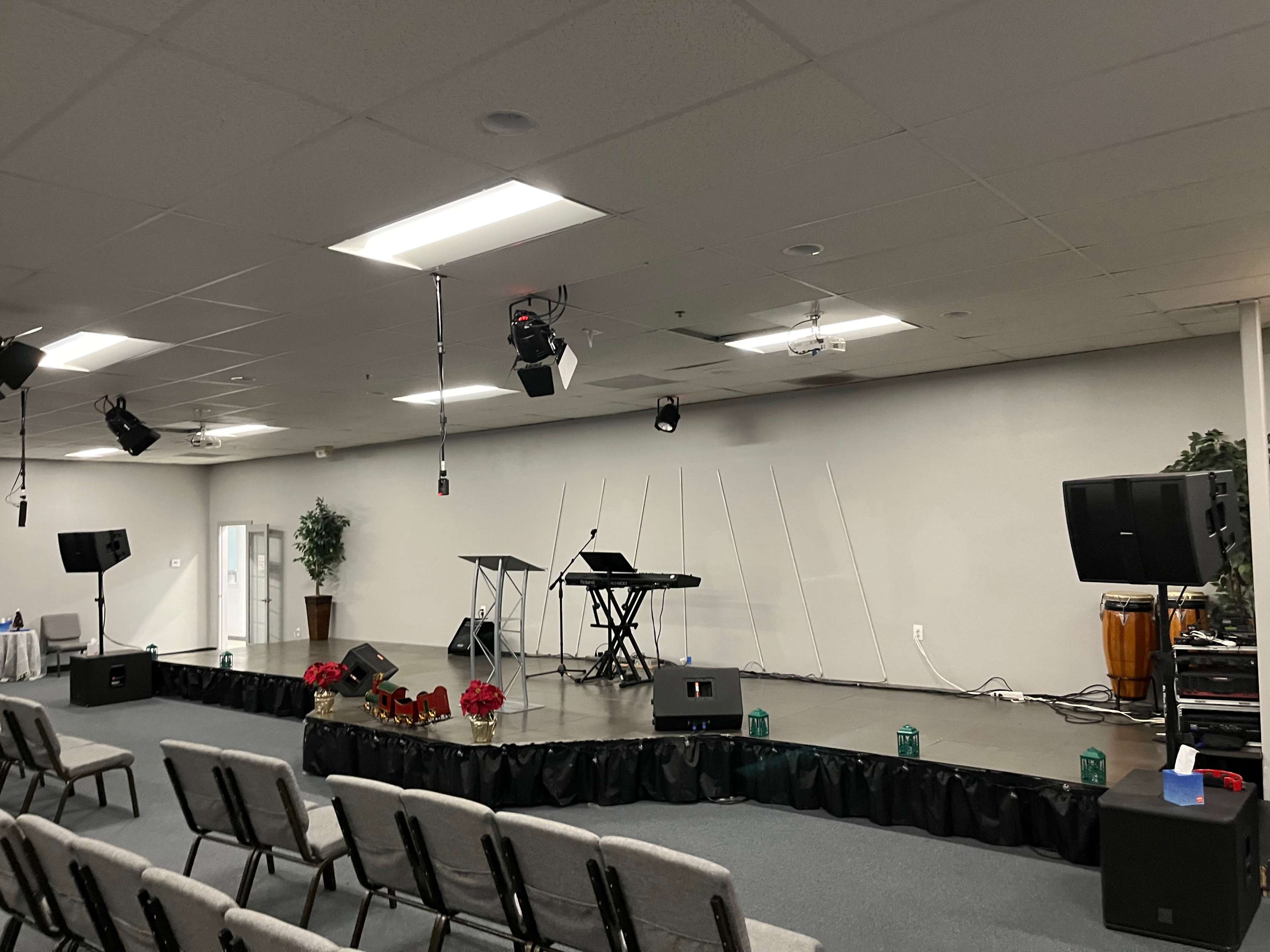 A stage set up in a church with seating in front, featuring a podium, keyboard, speakers, and decorative plants.