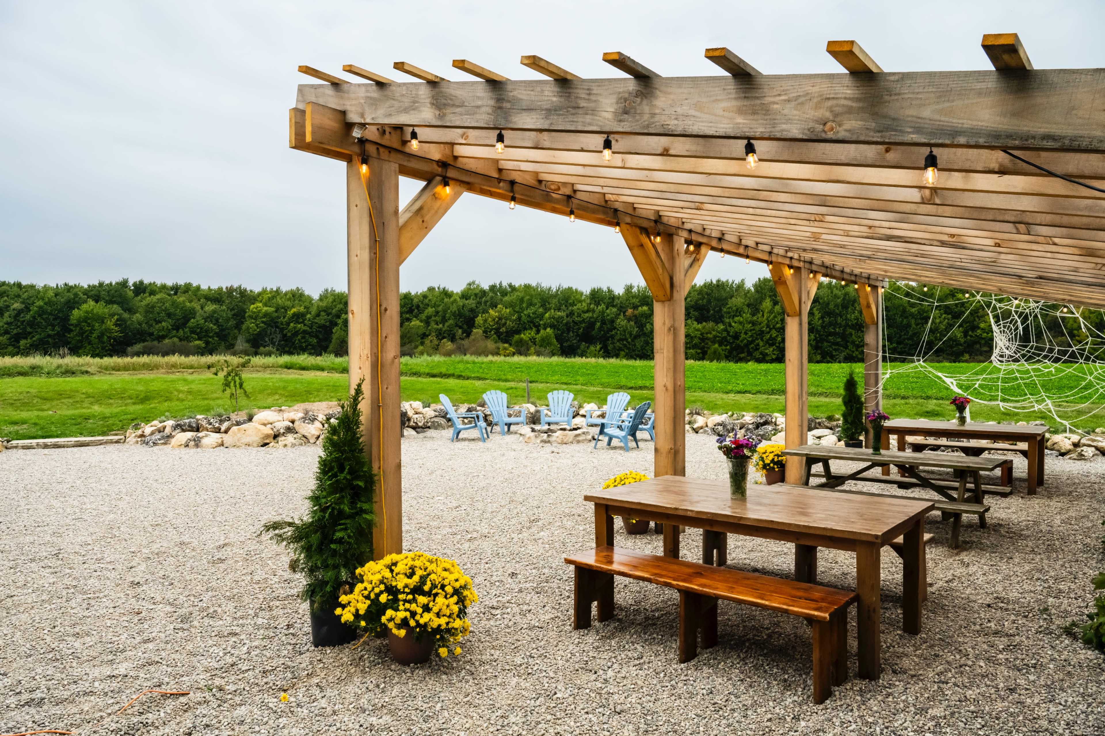 A wooden pergola with tables and chairs overlooks a green field, surrounded by gravel and decorated with potted plants.