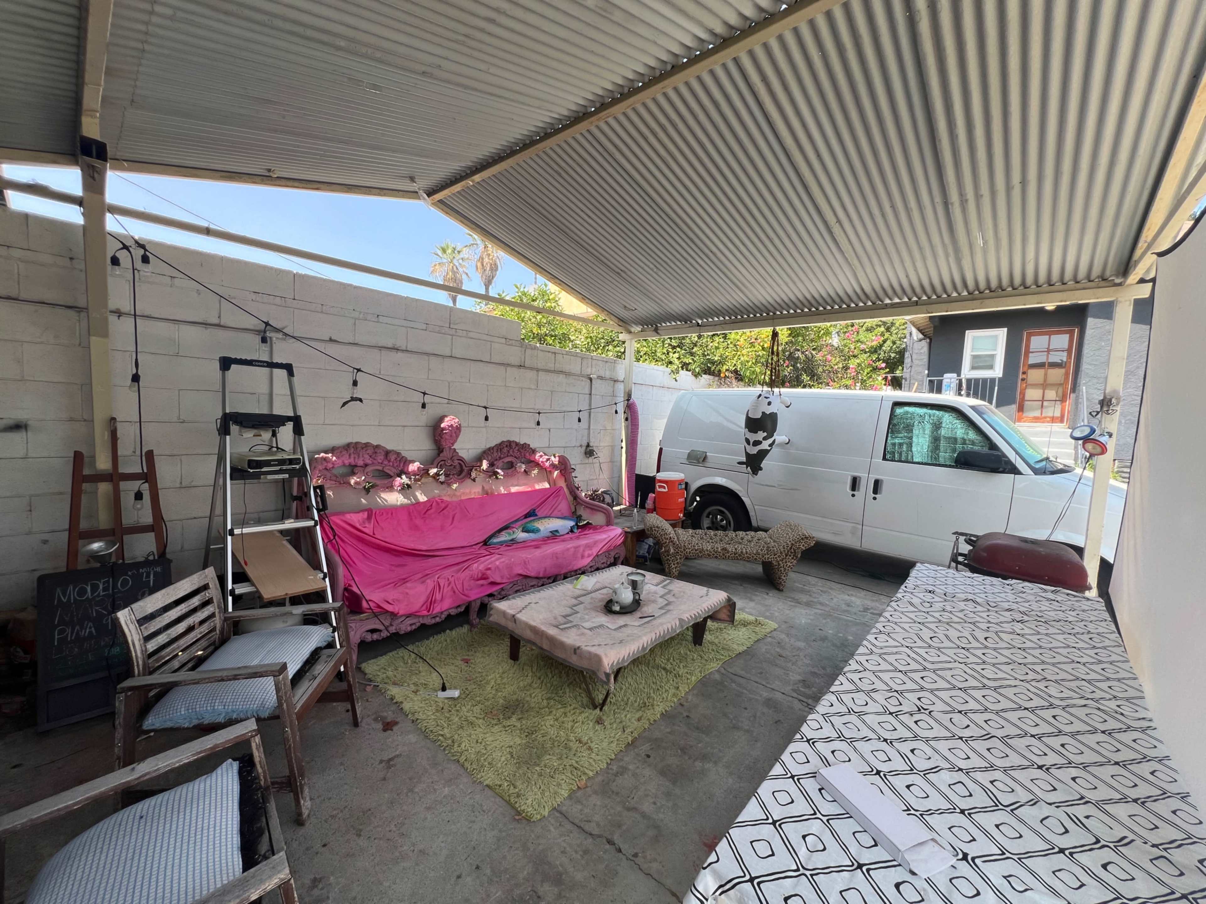 The image shows a covered outdoor space featuring a pink sofa, a couple of wooden chairs, a table, and a white van parked nearby.