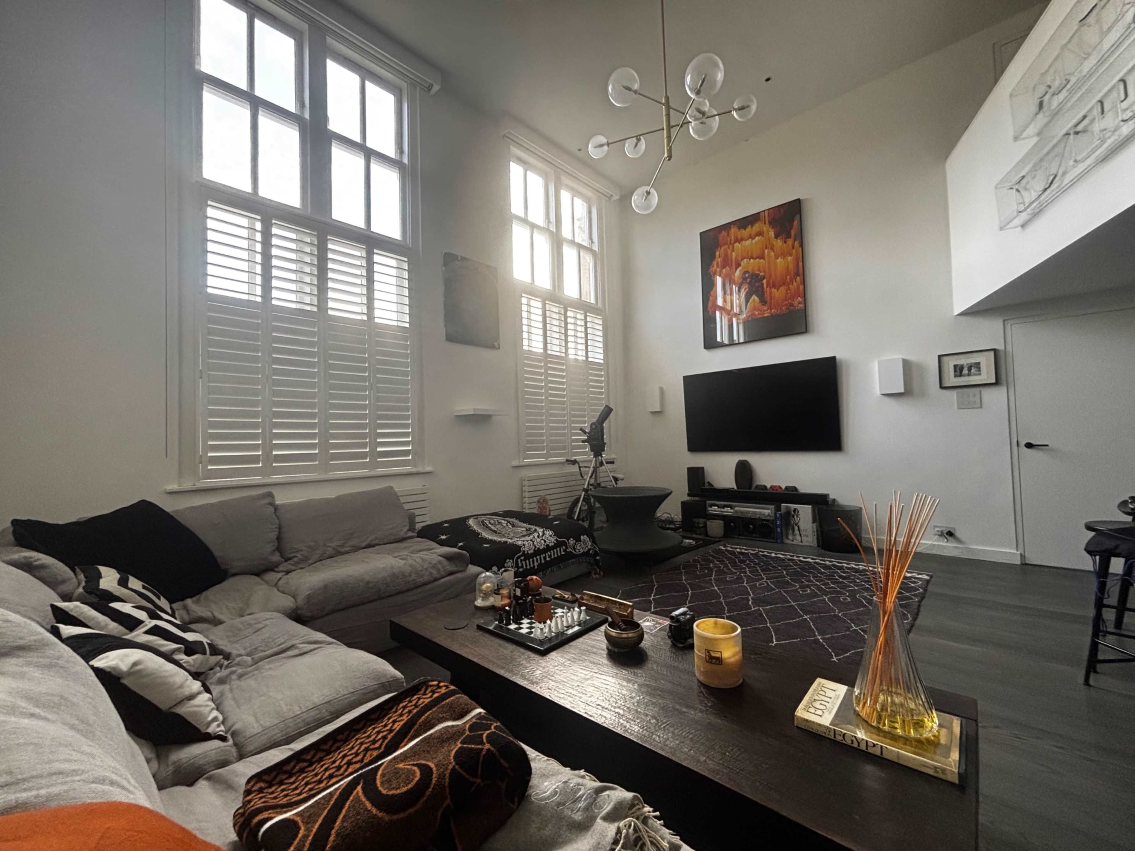The living room features a large sectional sofa, a coffee table with decorative items, a wall-mounted TV, and large windows with shutters, illuminated by a modern chandelier.