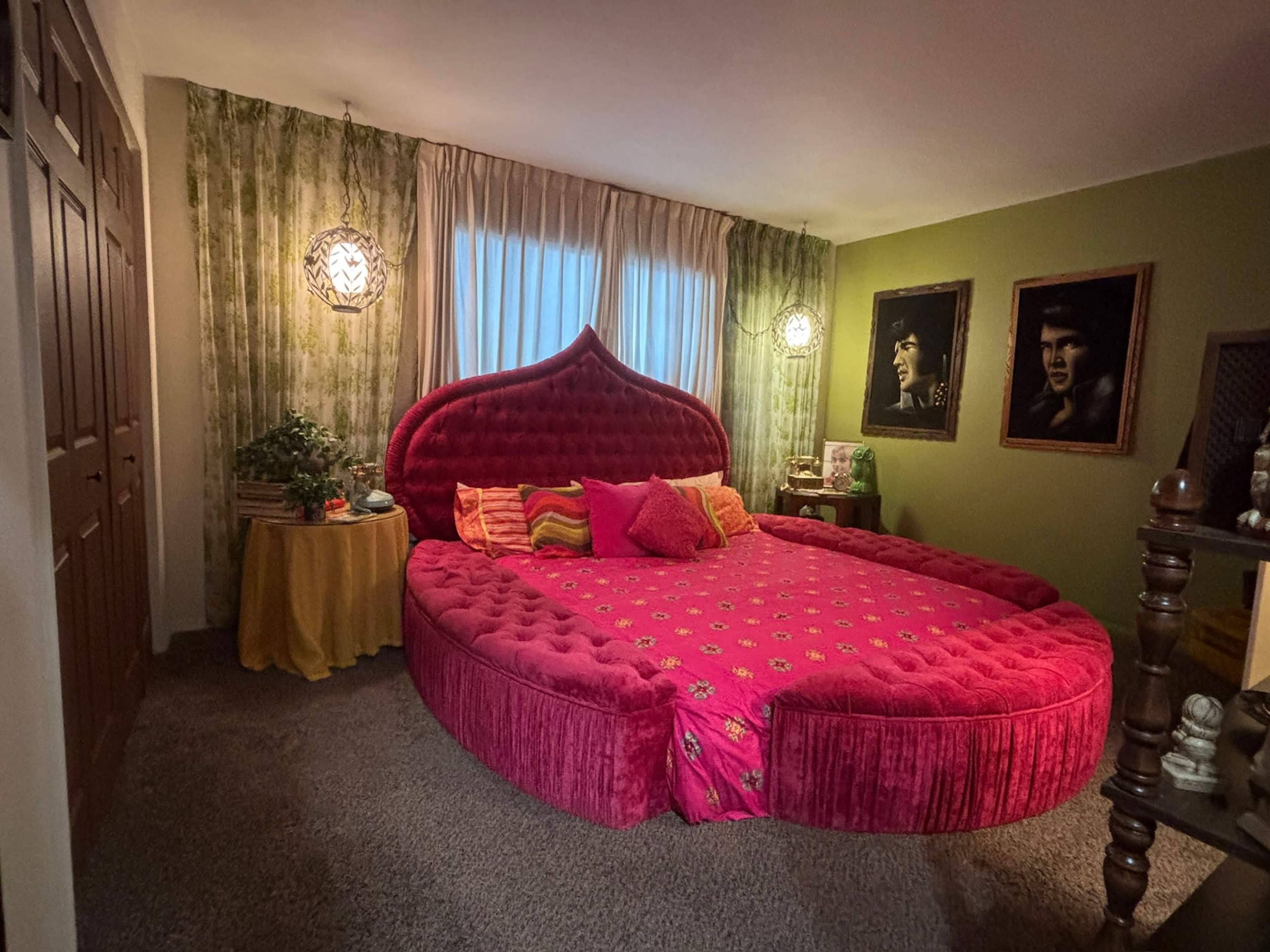 The image shows a uniquely shaped pink circular bed surrounded by green floral wallpaper, with two portraits on the wall and decorative lighting fixtures.