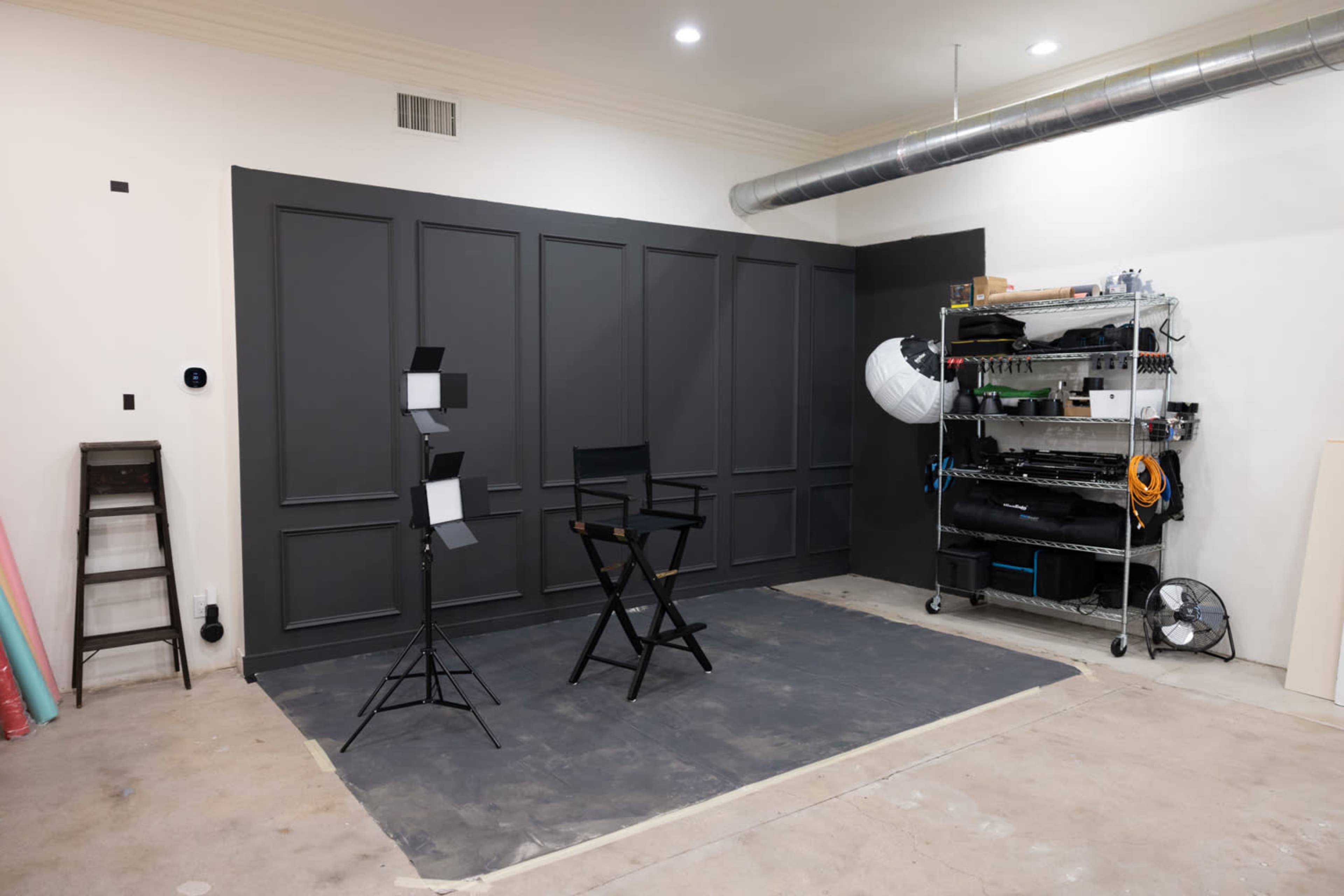A simple studio setup featuring a dark wall, a director's chair, two light stands, and a shelving unit containing various equipment.