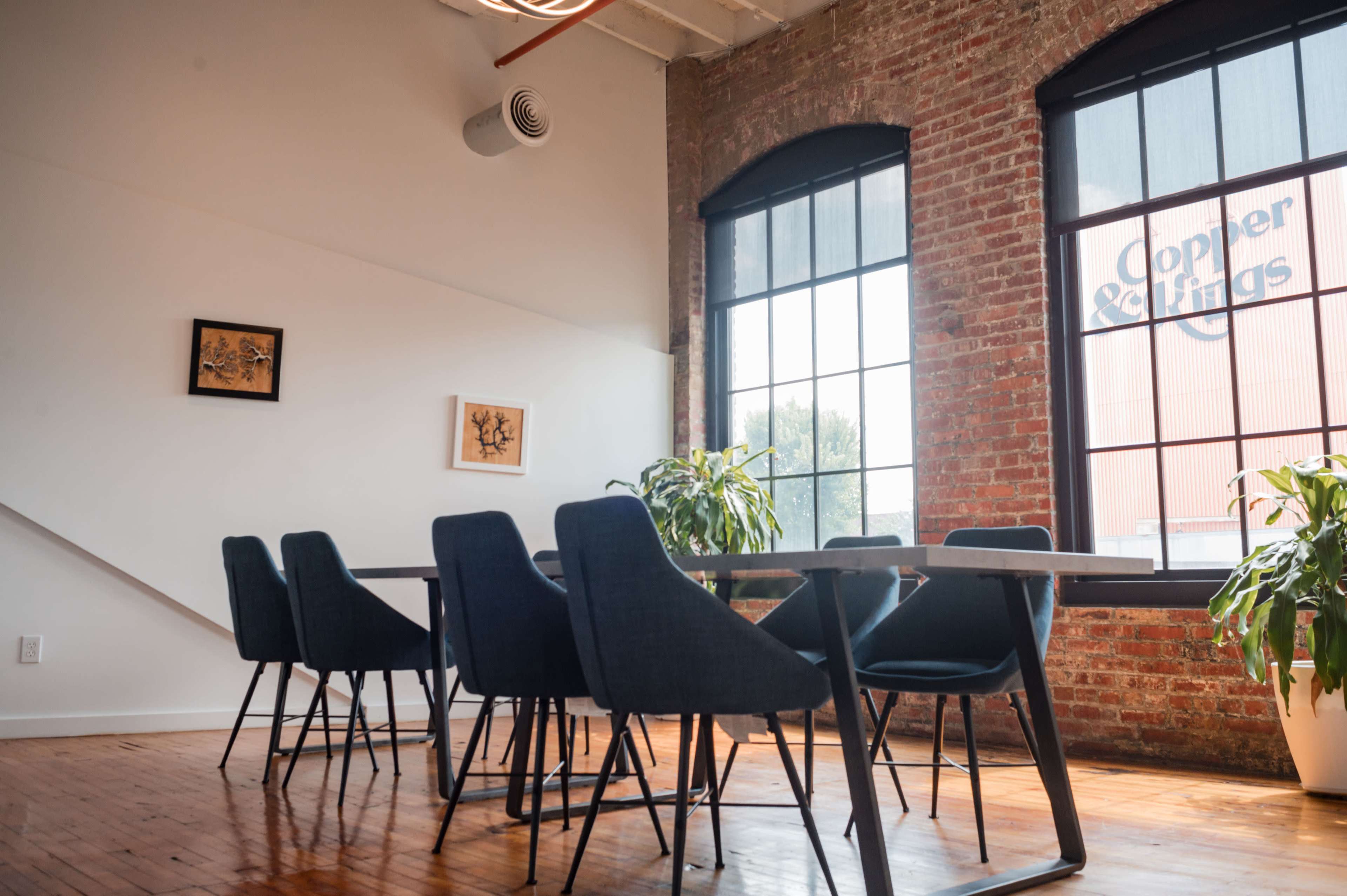 A modern conference room features a long table surrounded by dark chairs, with exposed brick walls and large windows revealing a view of a building outside.