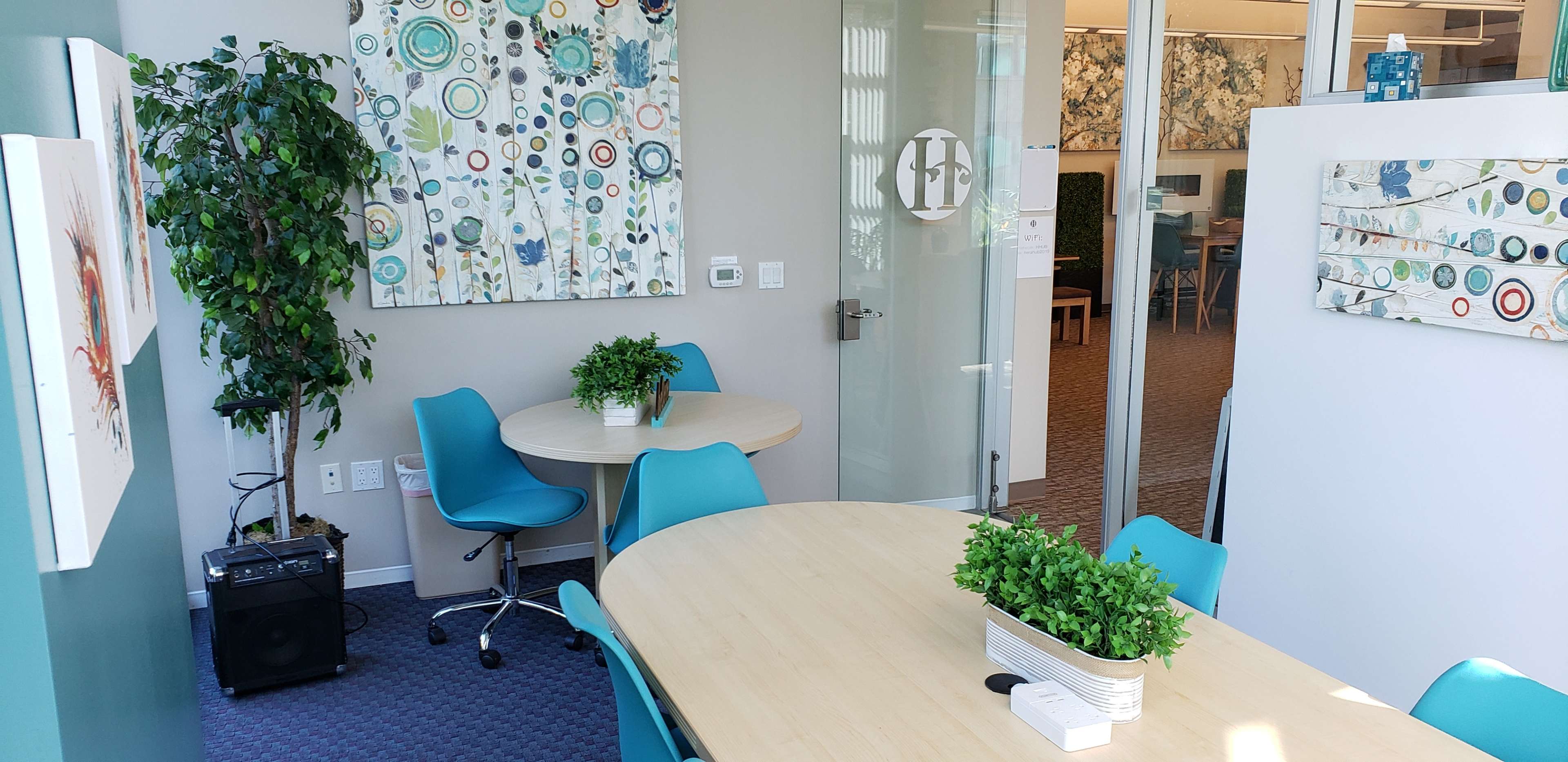 The image shows a modern office space featuring two round tables with teal chairs, decorative wall art, and a plant in a corner.
