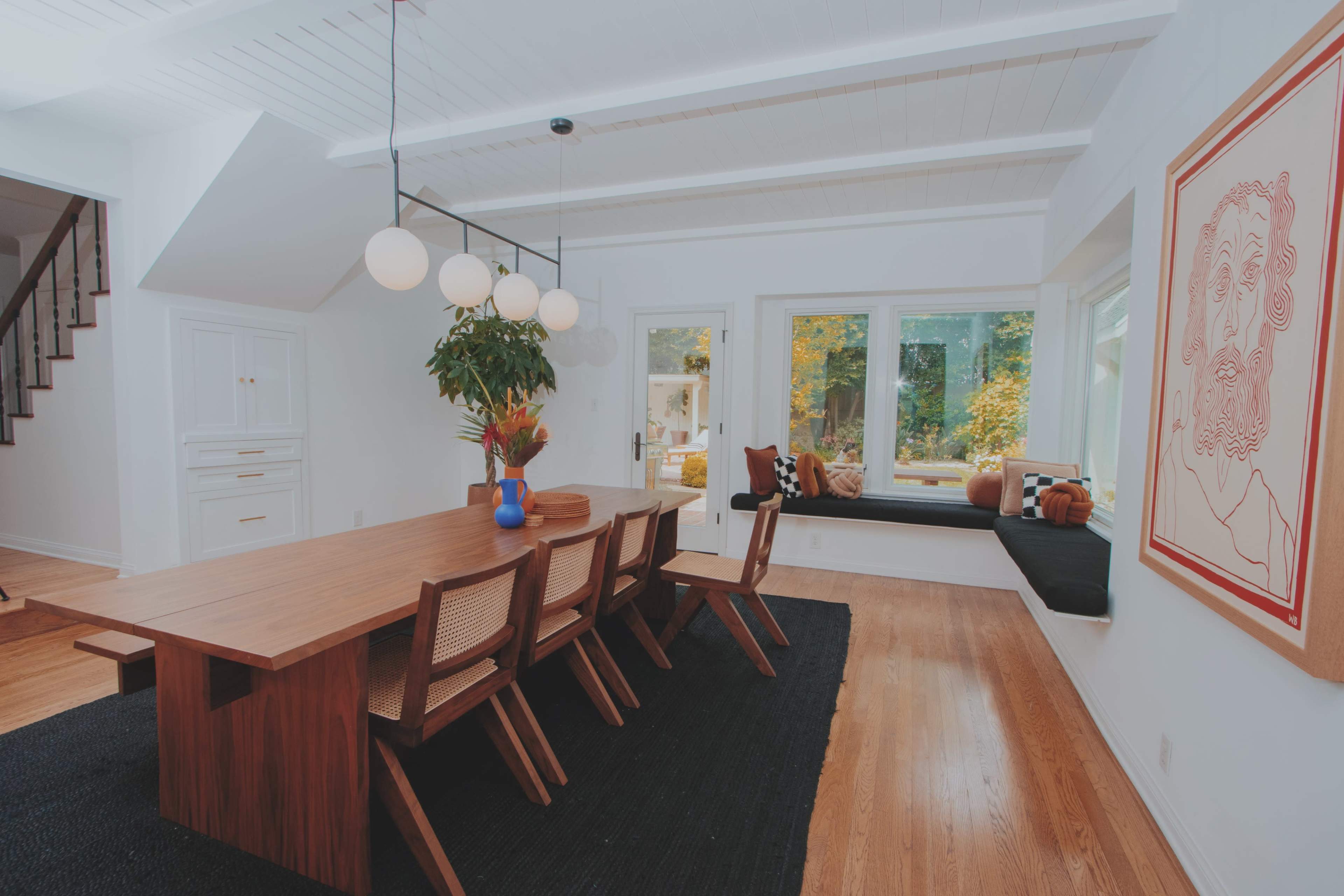 A modern dining room features a long wooden table with eight chairs, surrounded by large windows and a black rug.