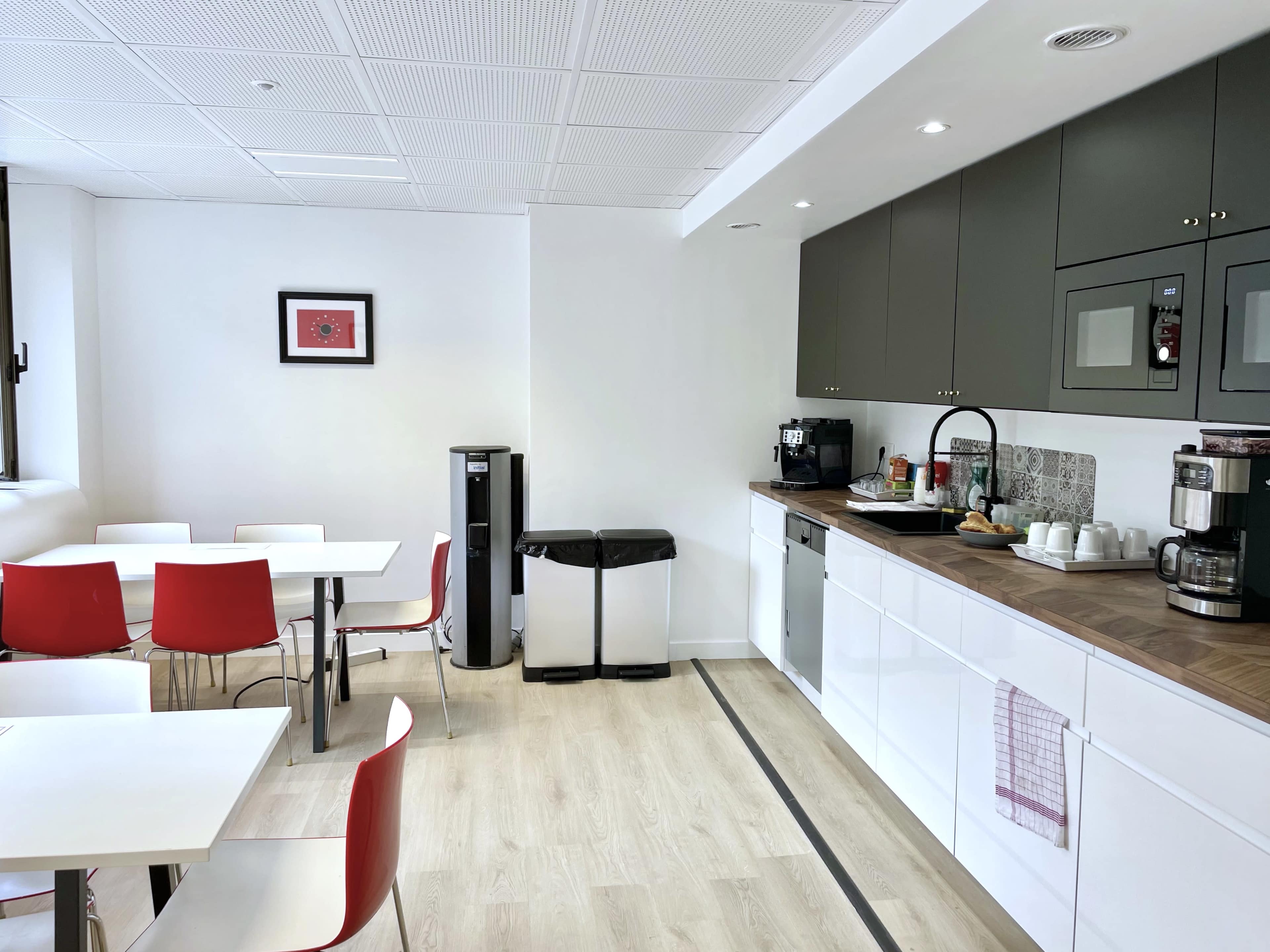 The image shows a modern break room featuring a kitchenette with cabinets, a coffee maker, and a dining area with white tables and red chairs.