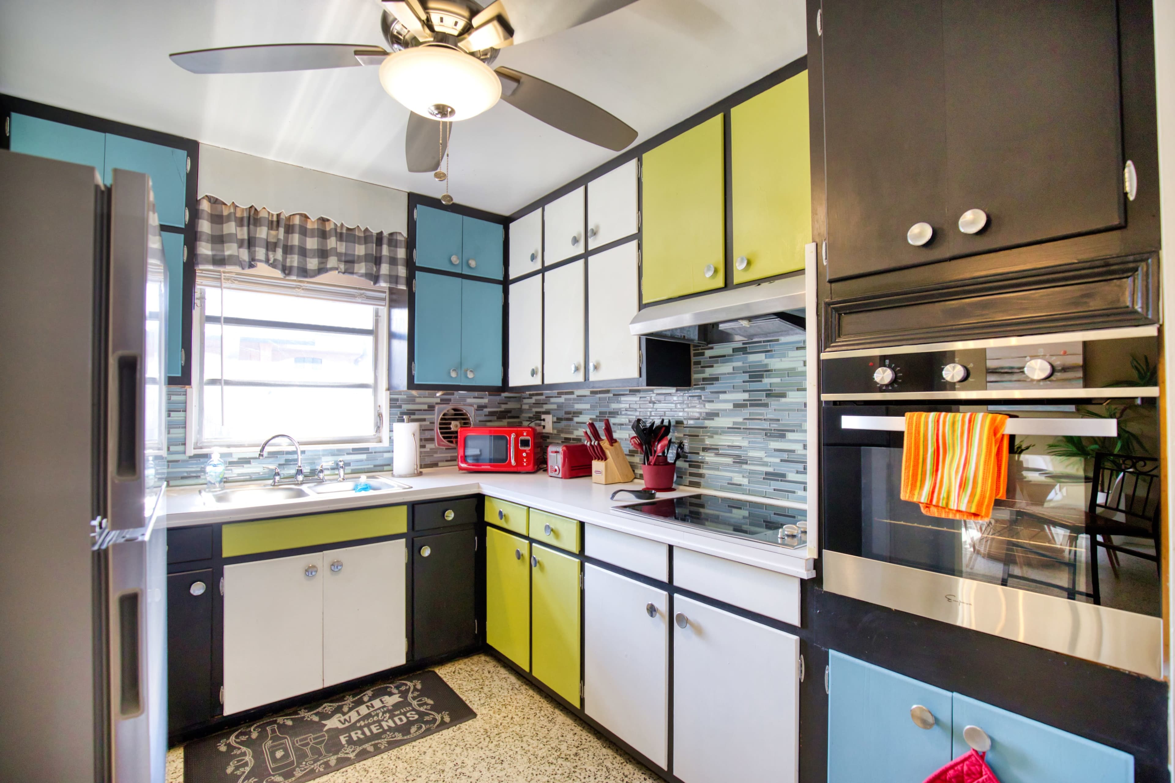 The kitchen features a mix of black, white, and brightly colored cabinets, with a window above the sink and various utensils arranged on the countertop.