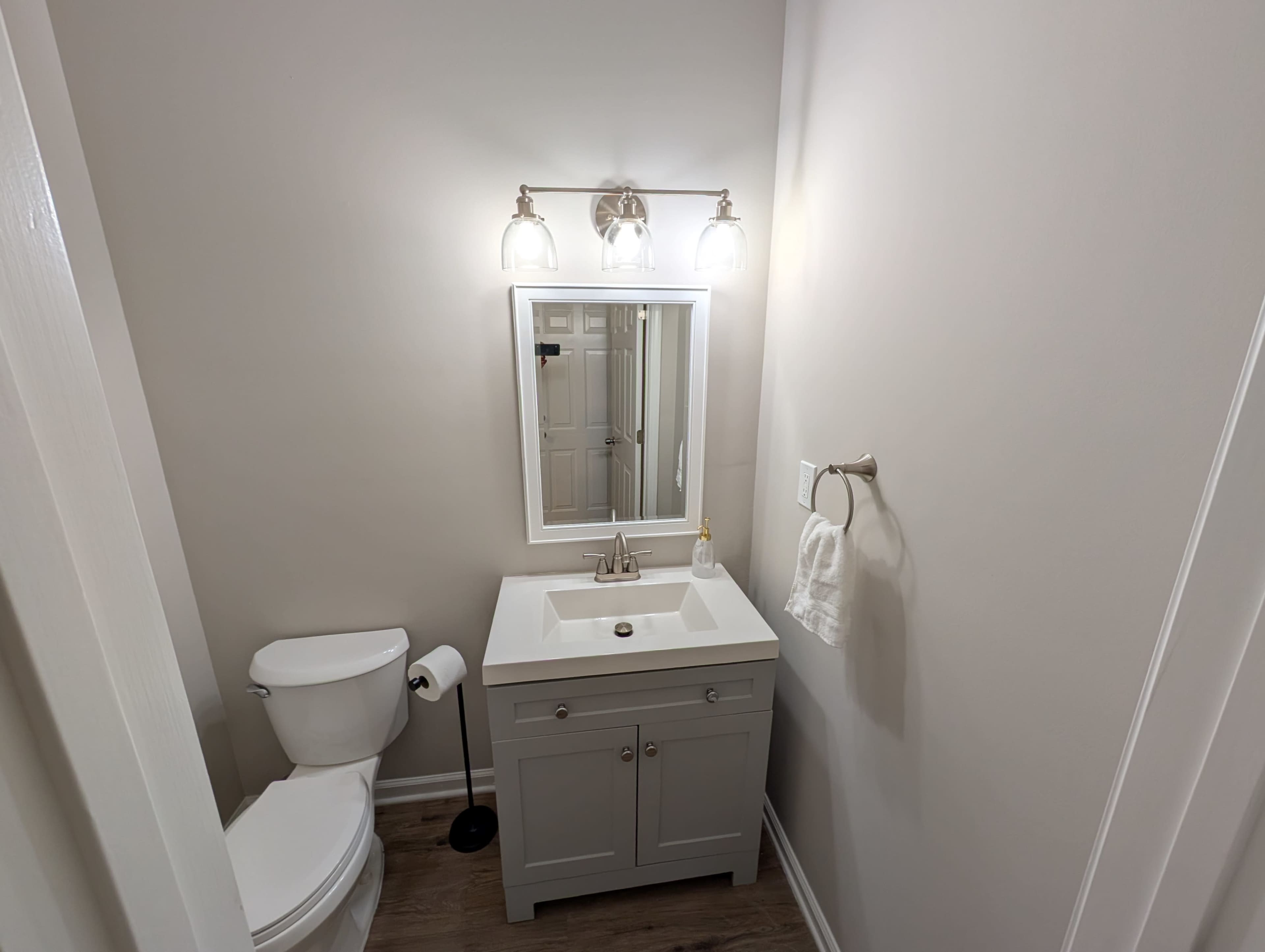 The image shows a small bathroom featuring a white vanity with a sink, a wall-mounted mirror, and a toilet.