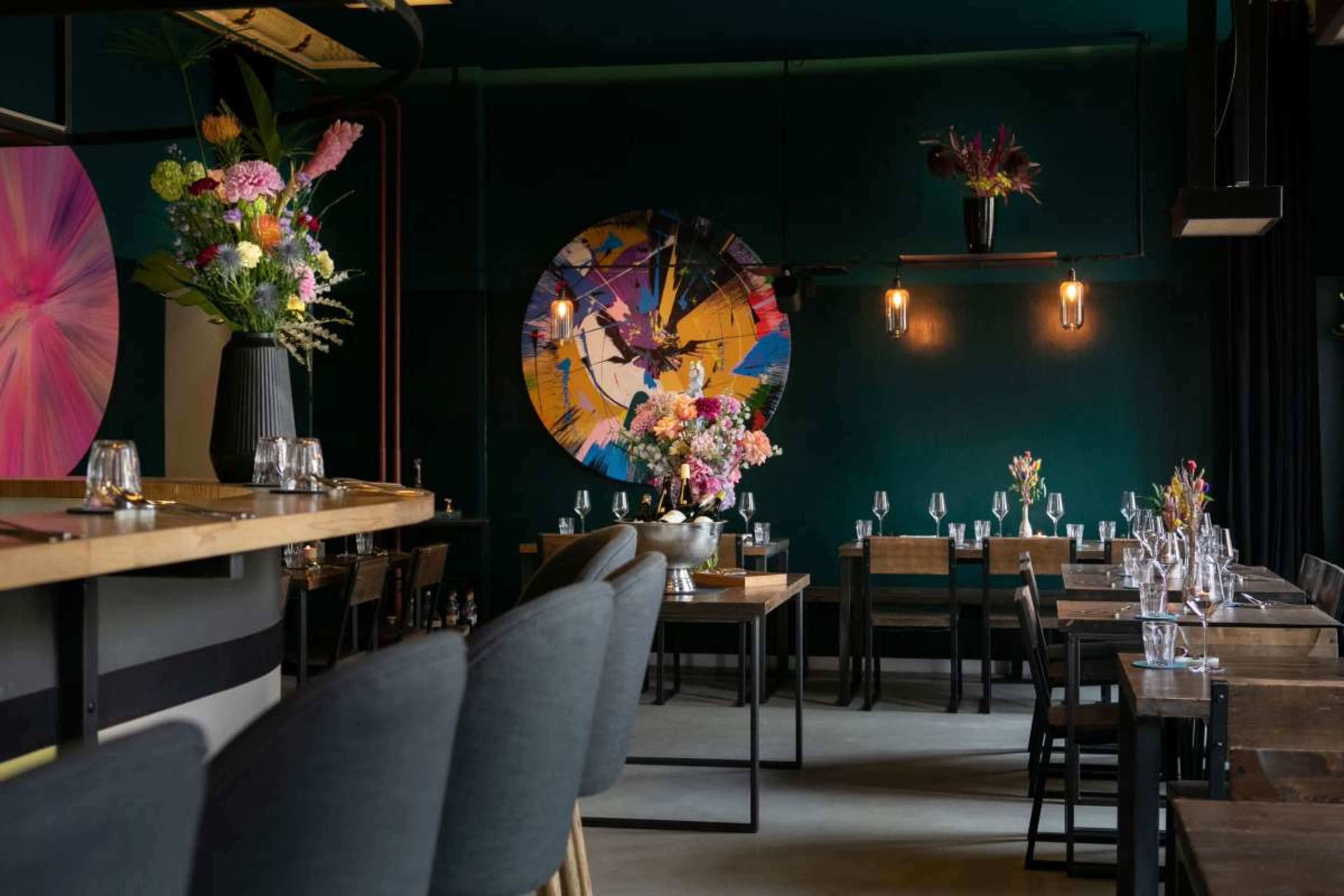 The image shows a stylish restaurant interior featuring wooden tables, dark-colored walls, and vibrant floral arrangements, complemented by abstract art on the walls.