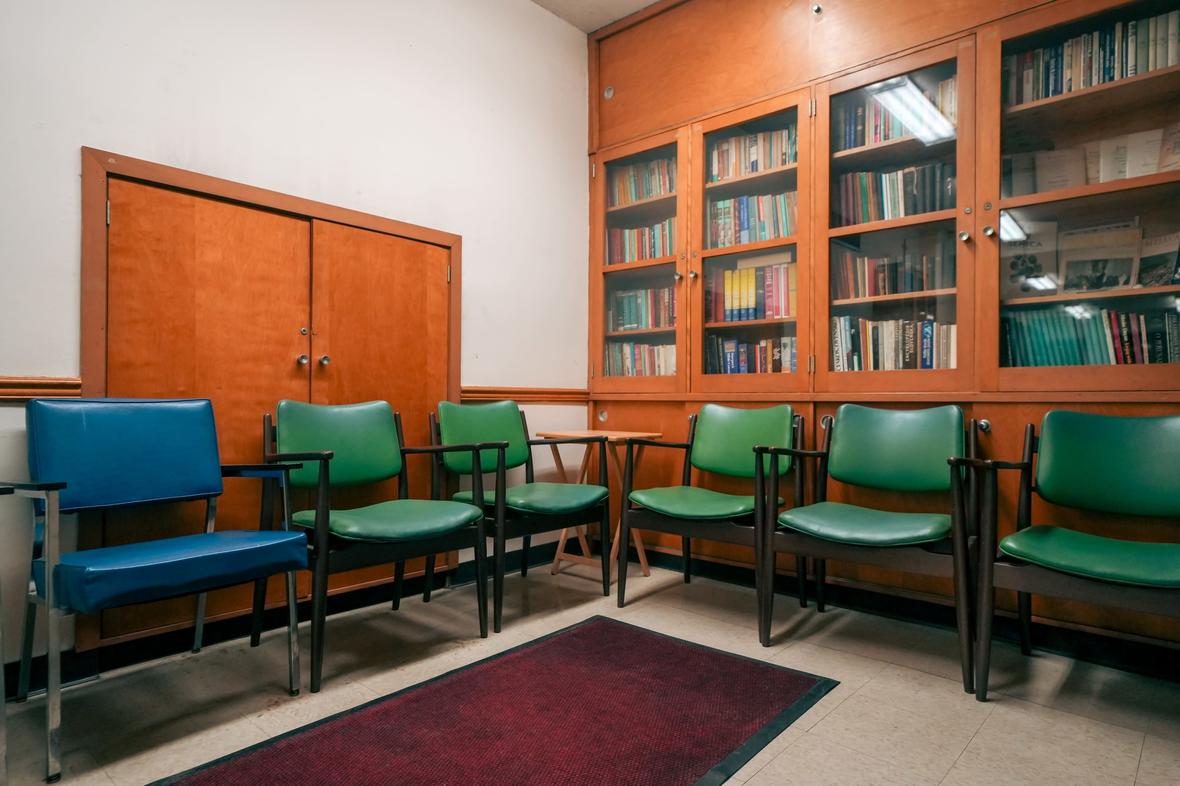 The image shows a waiting area with a row of green chairs, a single blue chair, and a wooden cabinet filled with books in the background.