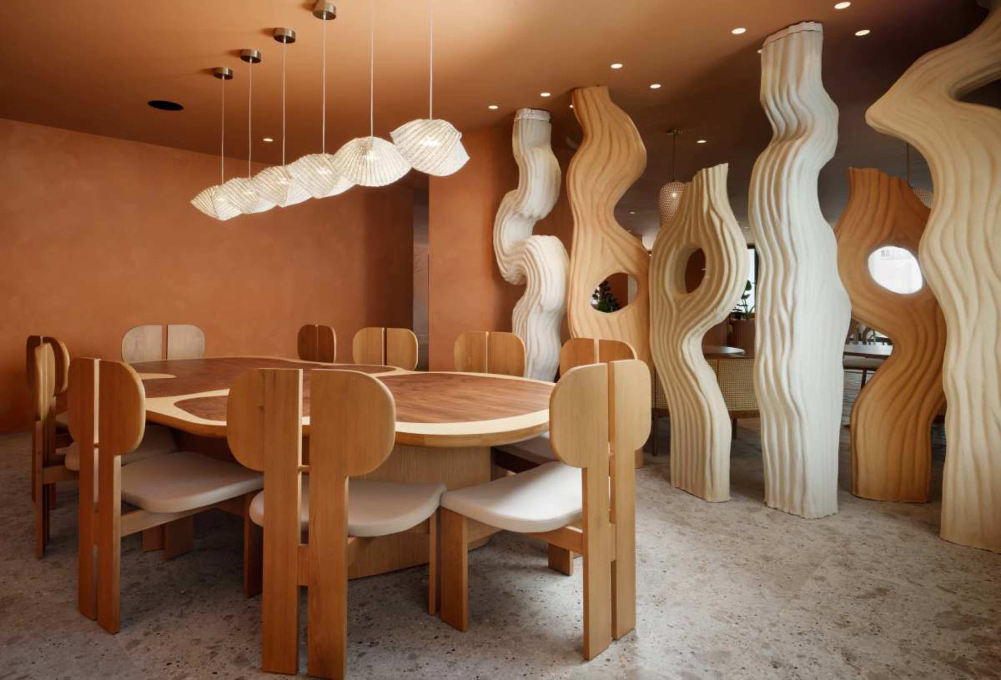 The image shows a modern dining room with a round wooden table surrounded by unique wooden chairs, illuminated by pendant lights, and accented by sculptural wall features.