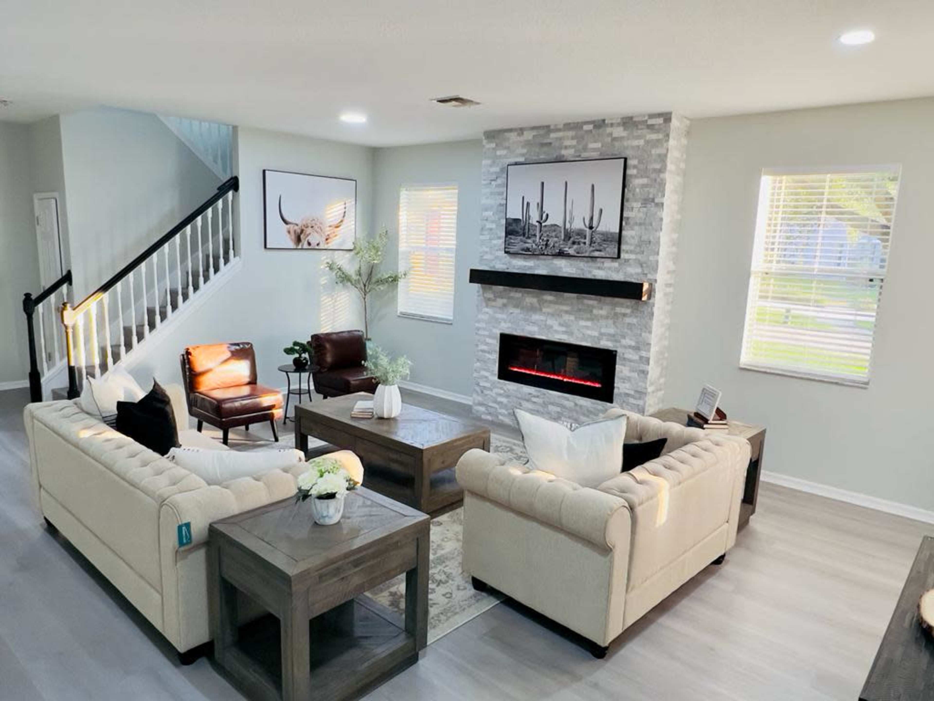 A modern living room featuring beige sofas, a stone fireplace, a coffee table, and a staircase in the background.