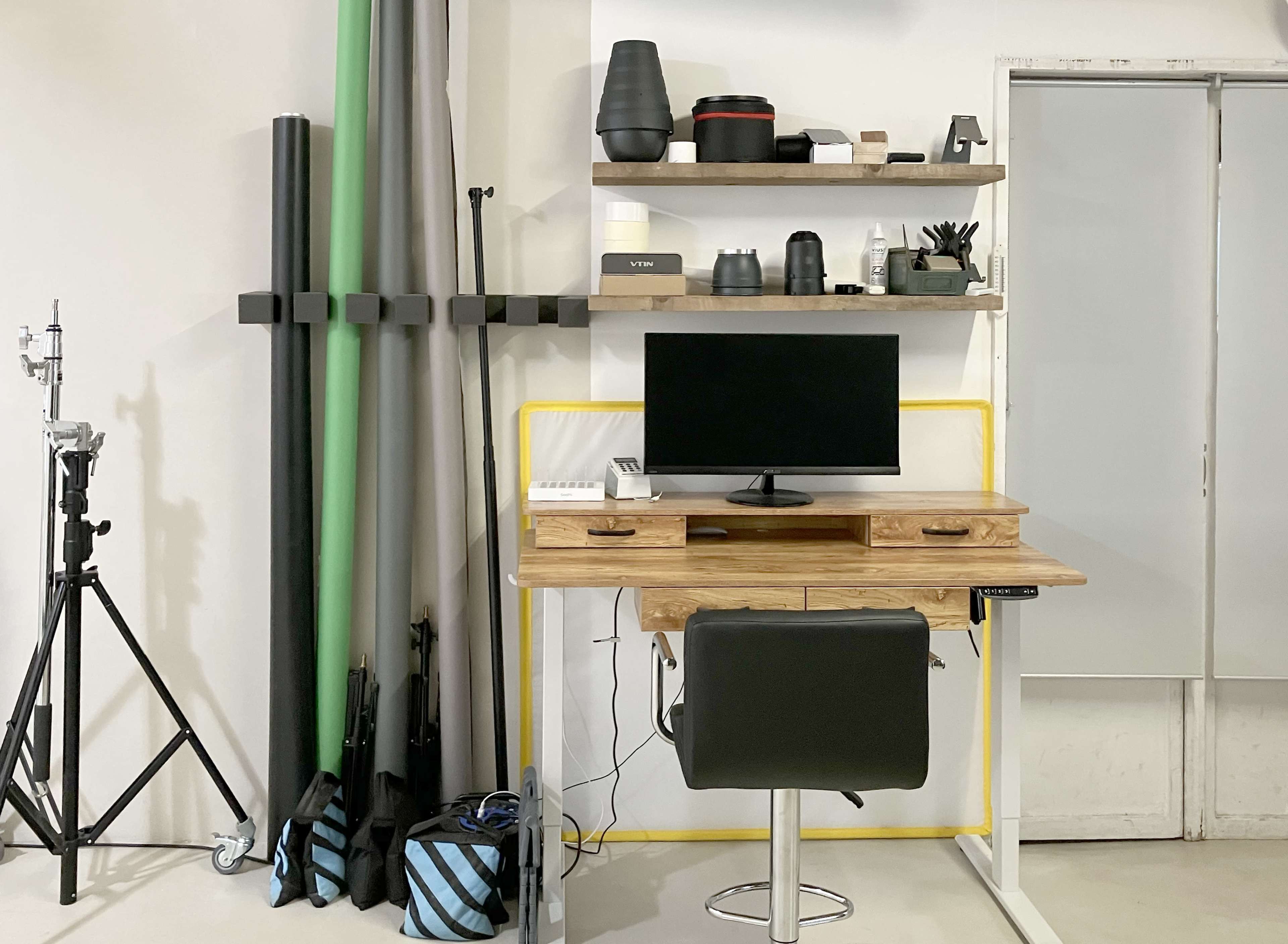A minimalist workspace with a wooden desk, a monitor on a stand, and various organizational items on shelves, alongside photography equipment arranged against a wall.