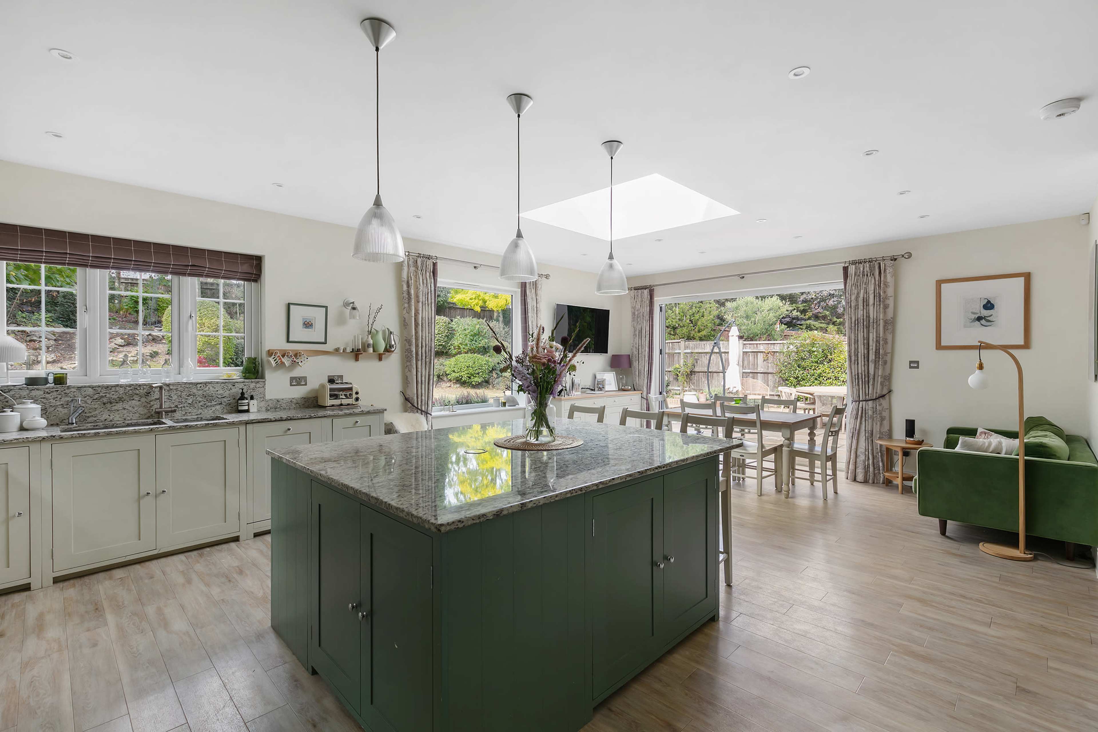 A spacious kitchen with green cabinetry features a large island and opens to a dining area with a table and chairs, illuminated by natural light from a skylight.