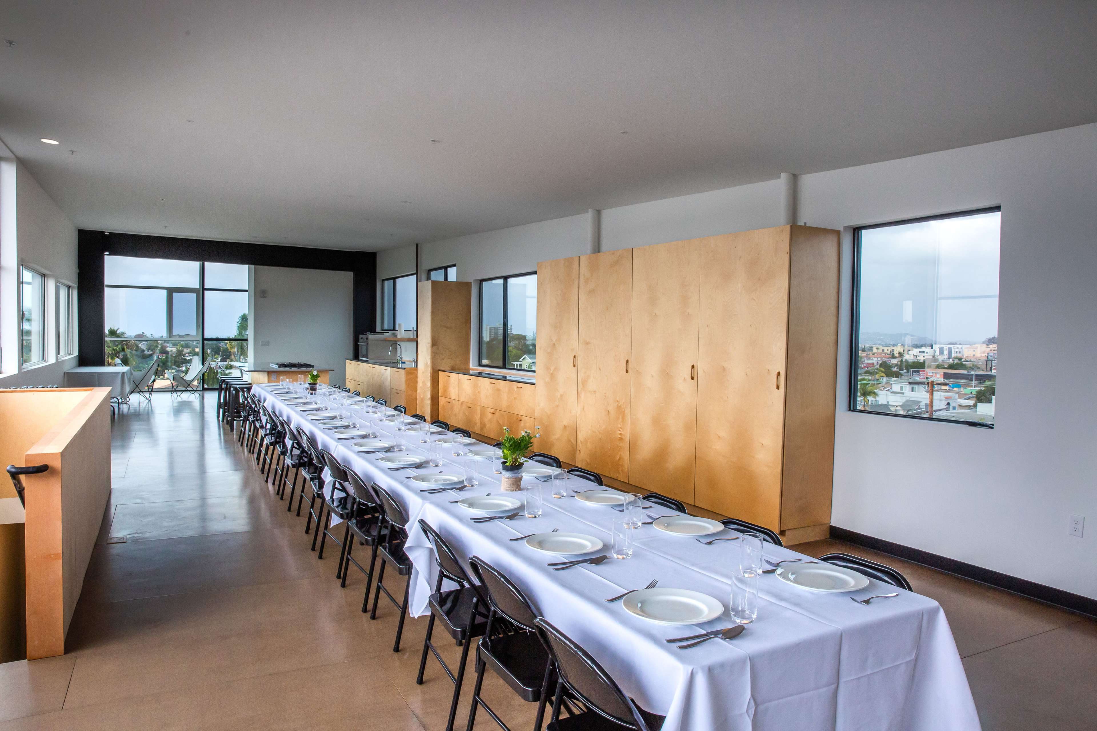 A long dining table is set with plates and glassware in a spacious room with large windows overlooking a landscape.