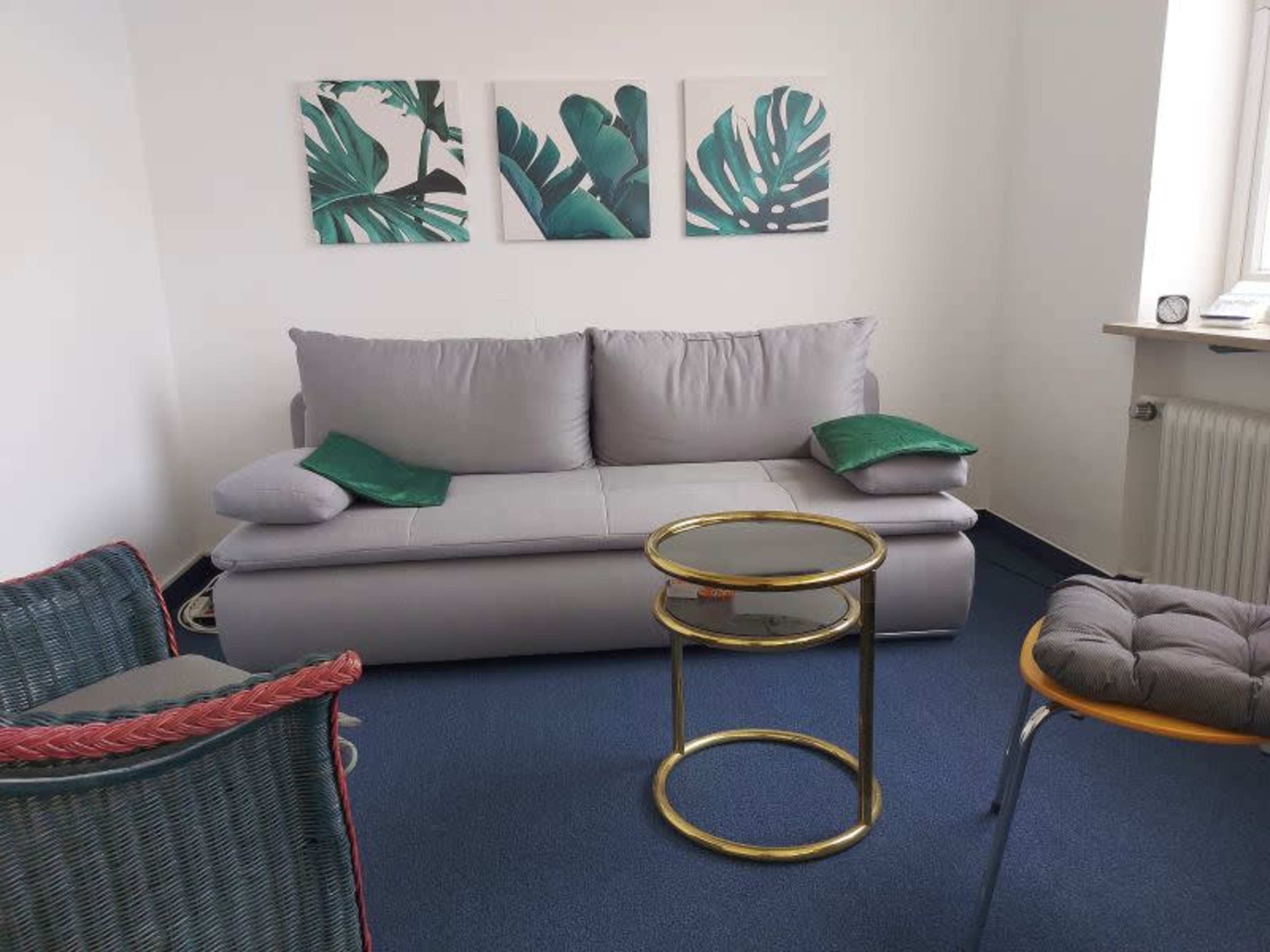 The image shows a small living space featuring a gray sofa with green cushions, a round glass coffee table, and two chairs, one of which is woven, along with three wall-mounted tropical leaf prints.