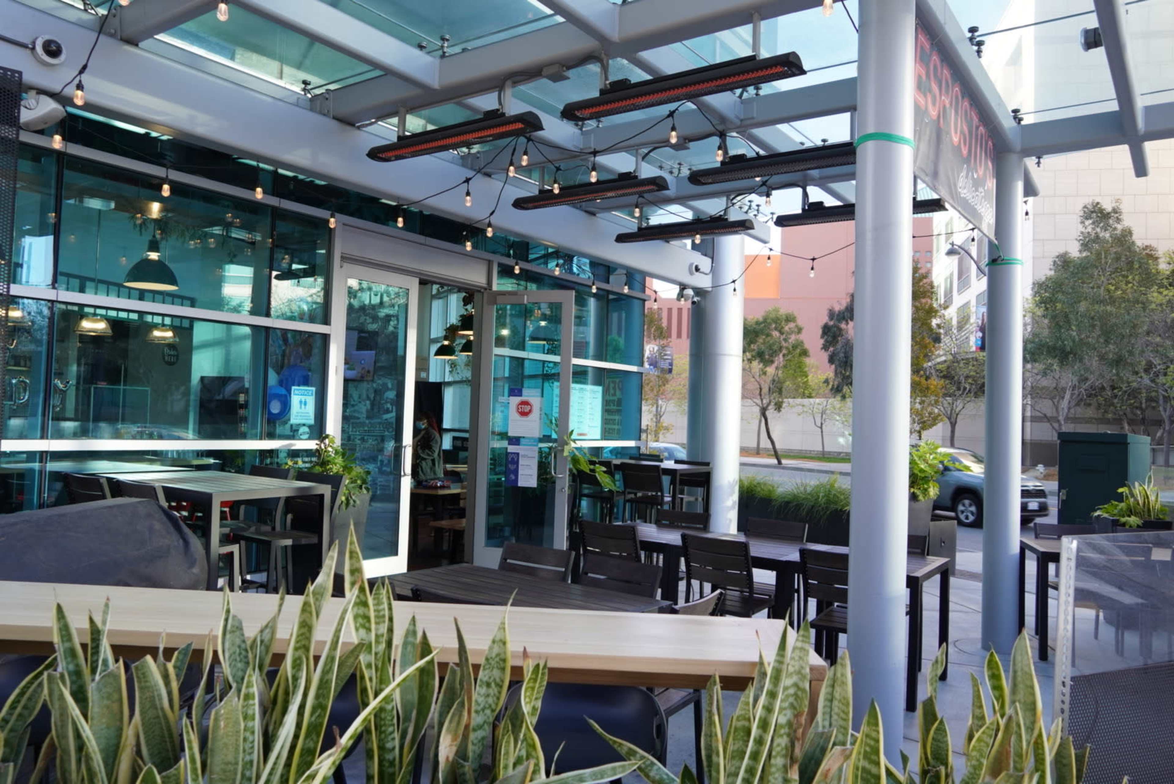 A modern outdoor dining area features tables, chairs, and hanging lights beneath a glass roof with greenery along the edges.