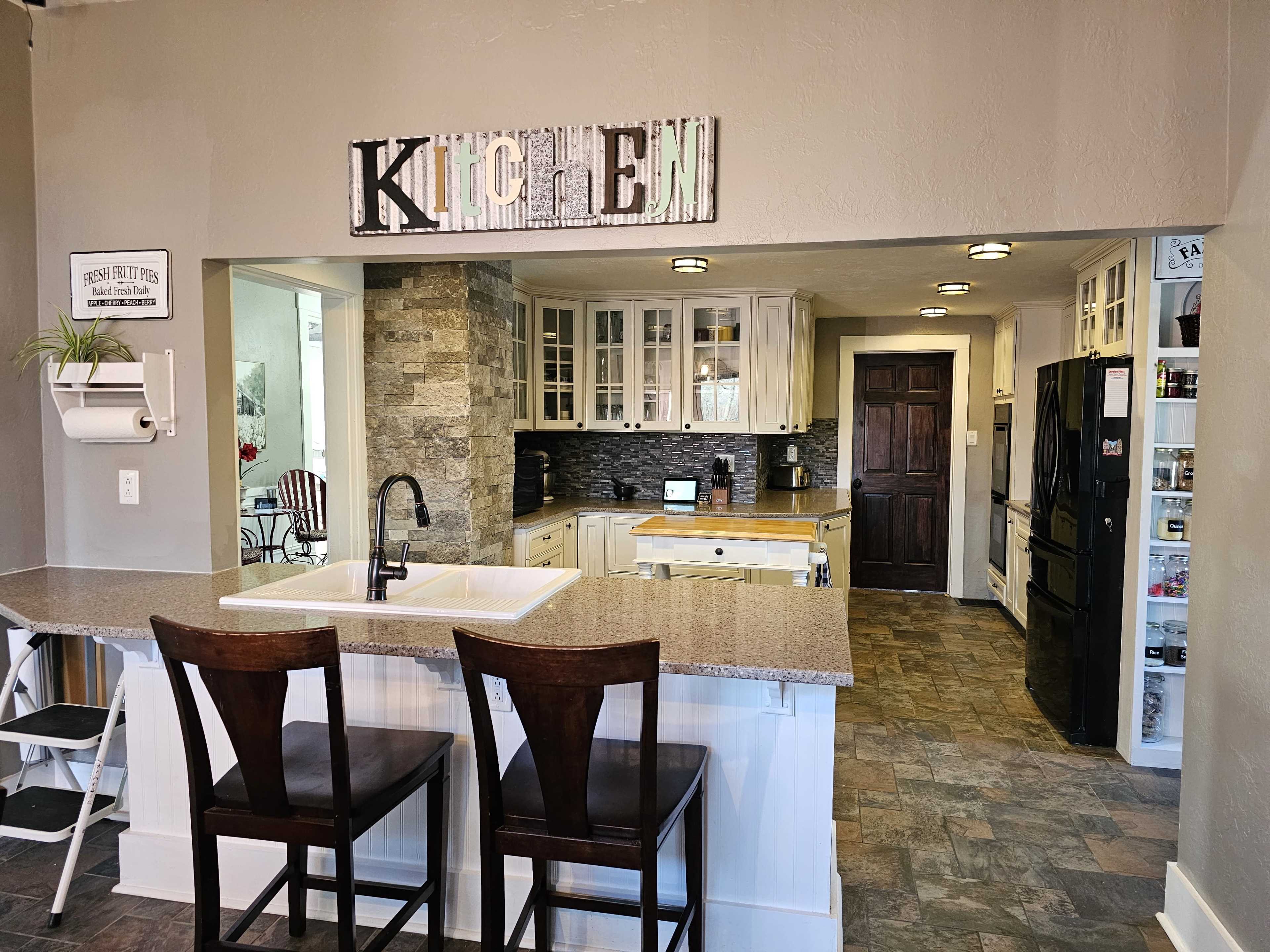 A kitchen features a central island with two bar stools, cabinetry, and a tiled backsplash, leading to a spacious cooking area with modern appliances.