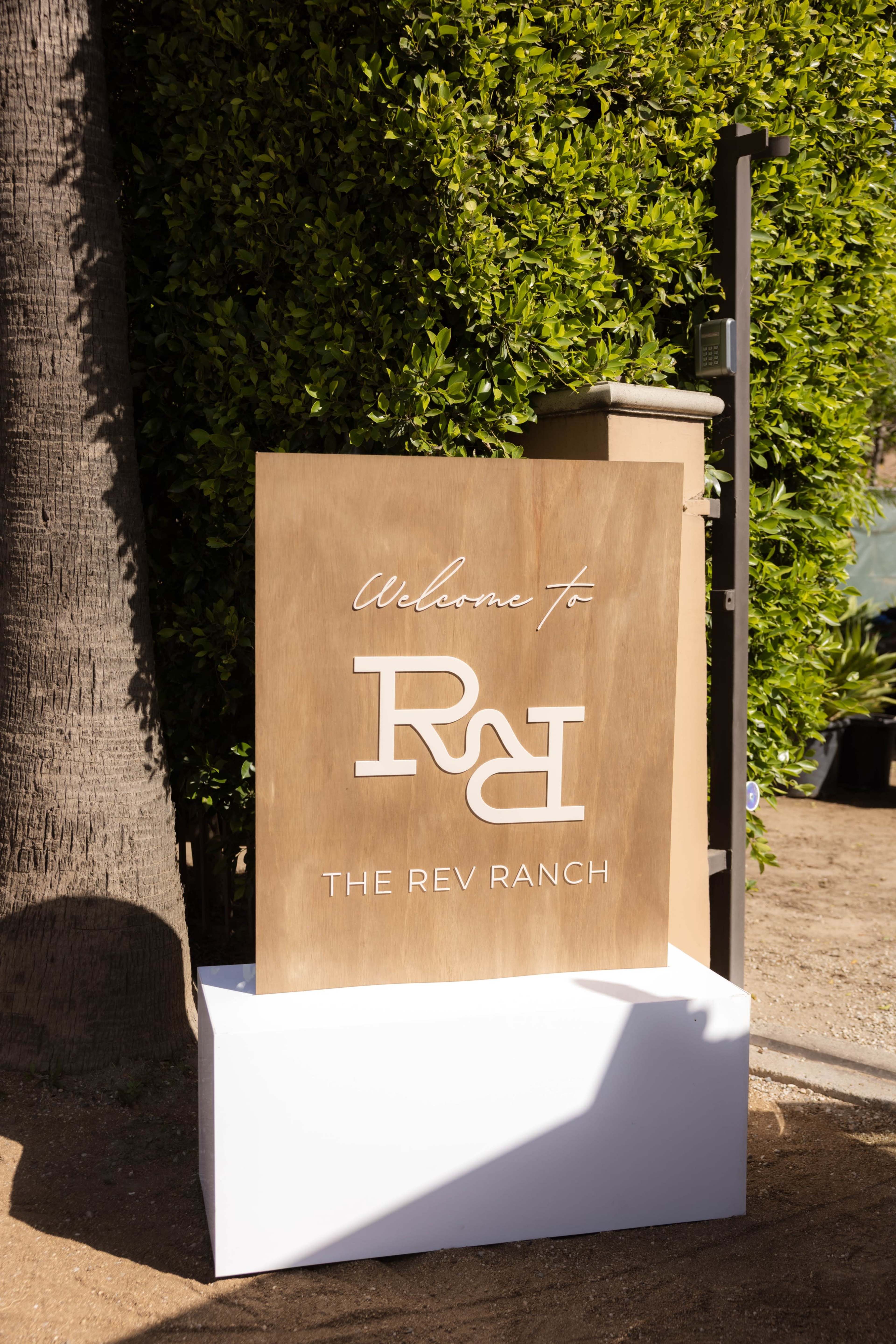 A wooden sign with the text "Welcome to The Rev Ranch" is positioned next to a green hedge.
