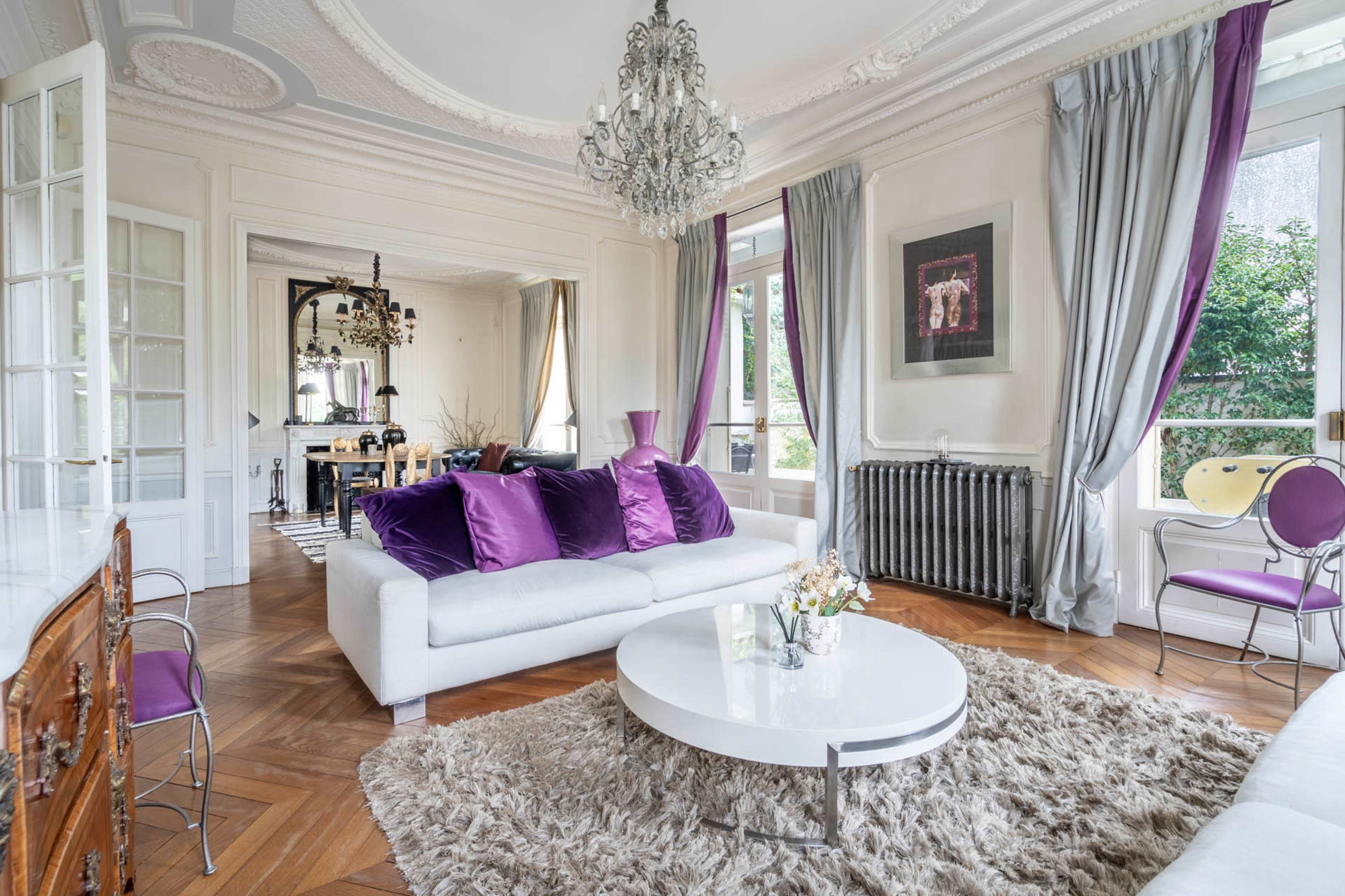 The image shows a stylish living room with a white sofa, purple accent pillows, a round coffee table, and large windows overlooking a garden.