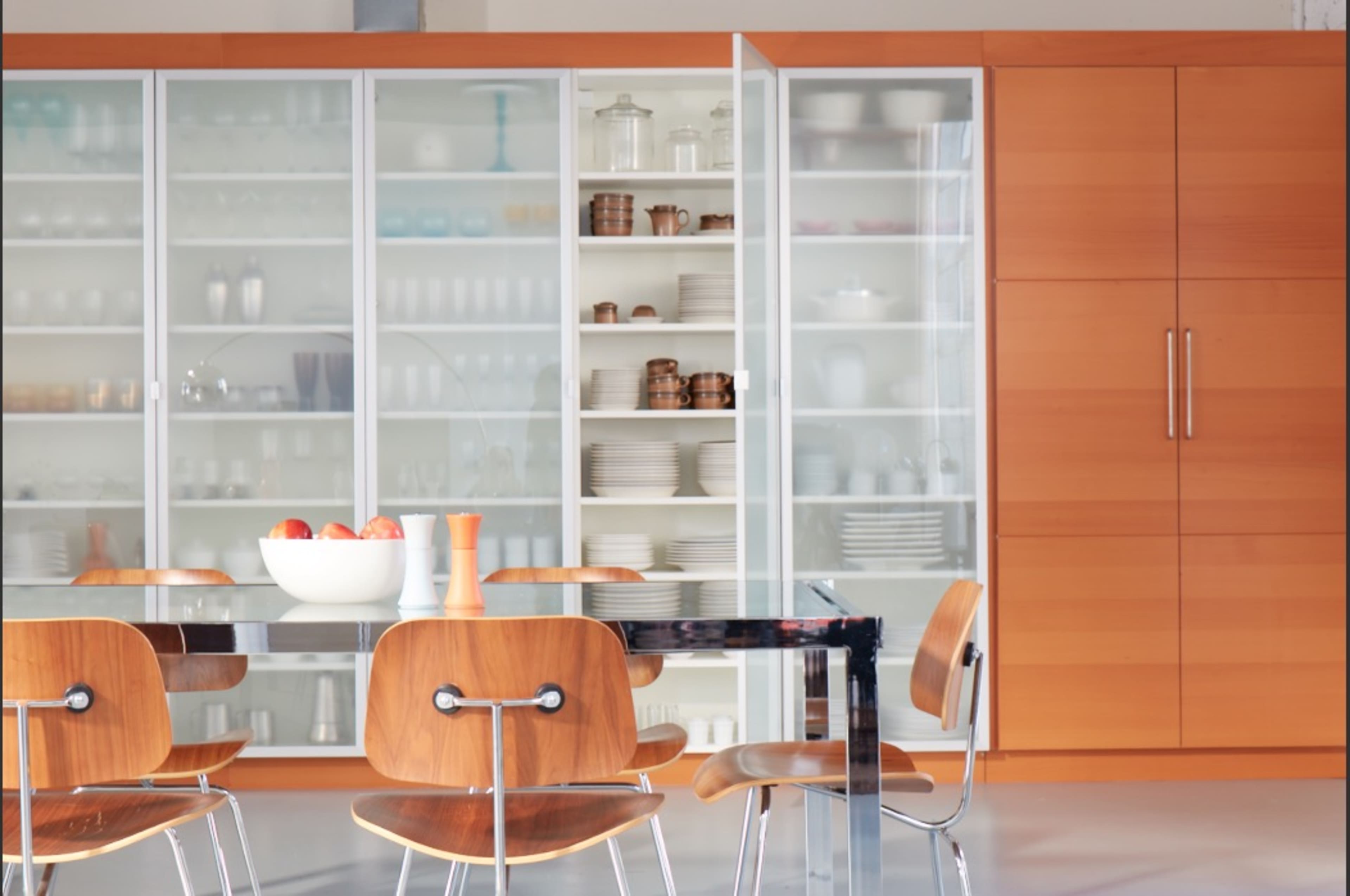 The image shows a modern dining area featuring a glass-top table surrounded by wooden chairs, with a large cabinet of glass doors displaying dishware and kitchen items.