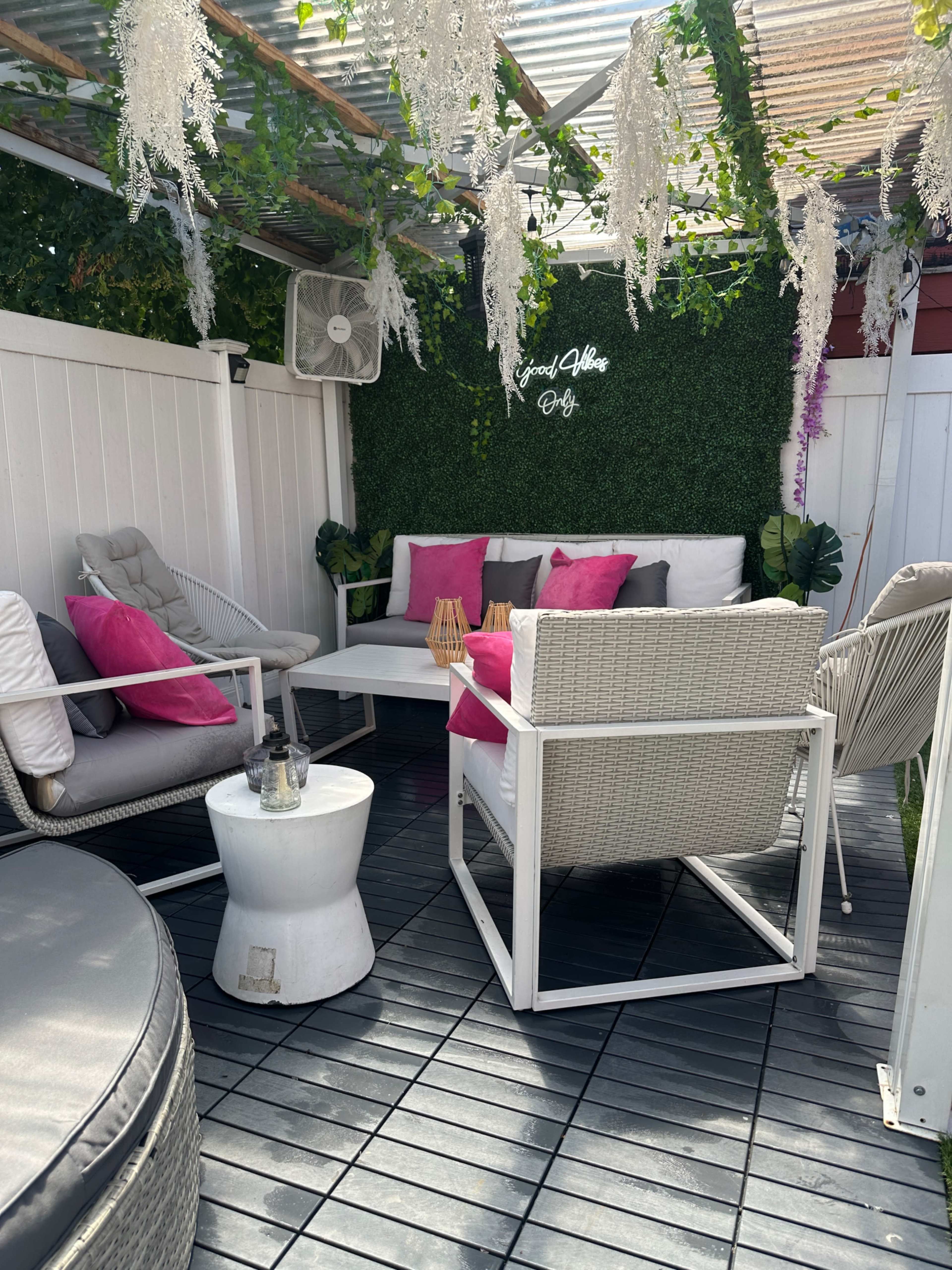 A cozy outdoor seating area with white furniture, pink cushions, and decorative greenery on the walls.