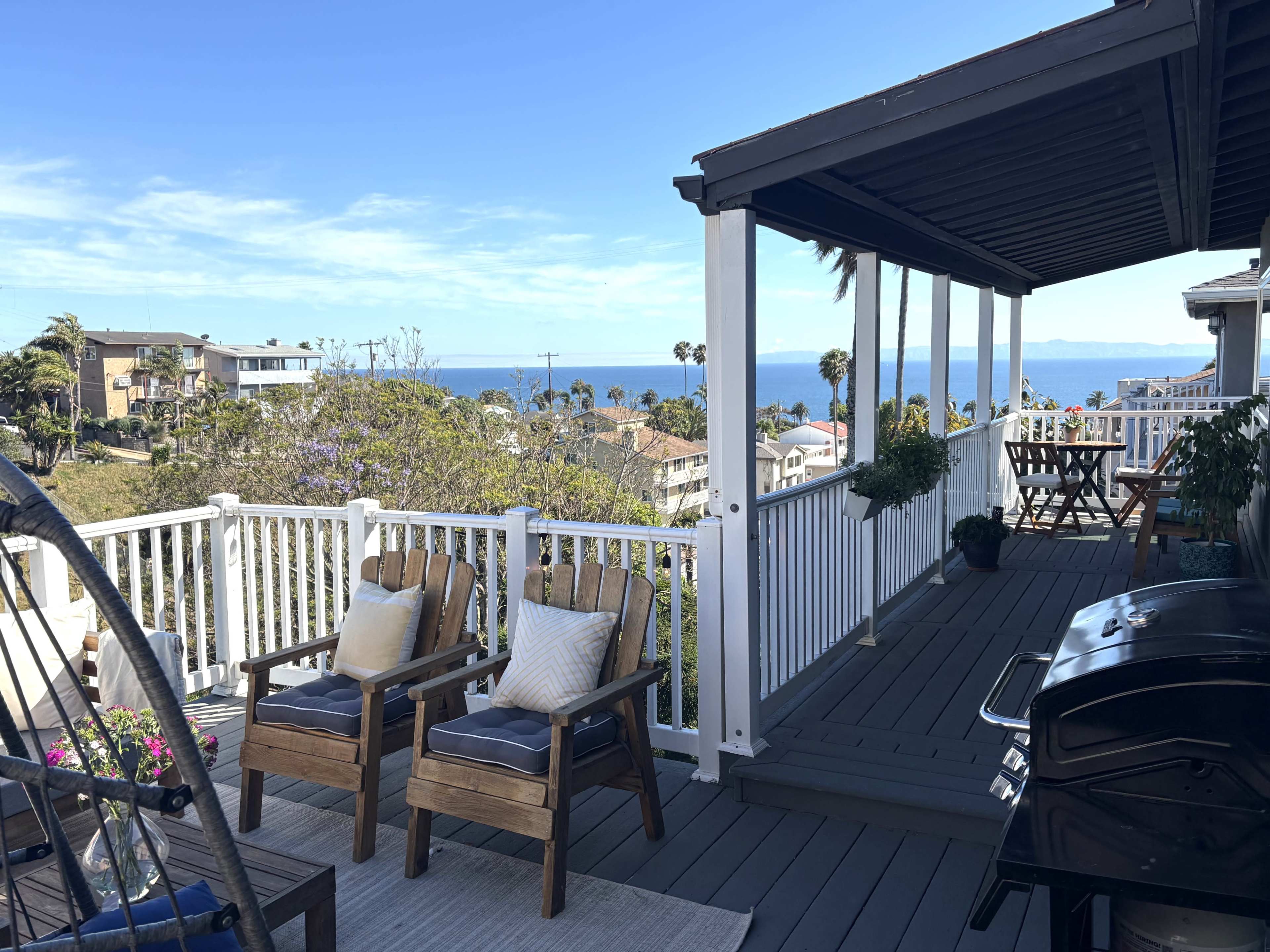 The image shows a spacious deck with wooden furniture, a barbecue grill, and a view of the ocean and coastal houses under a clear blue sky.
