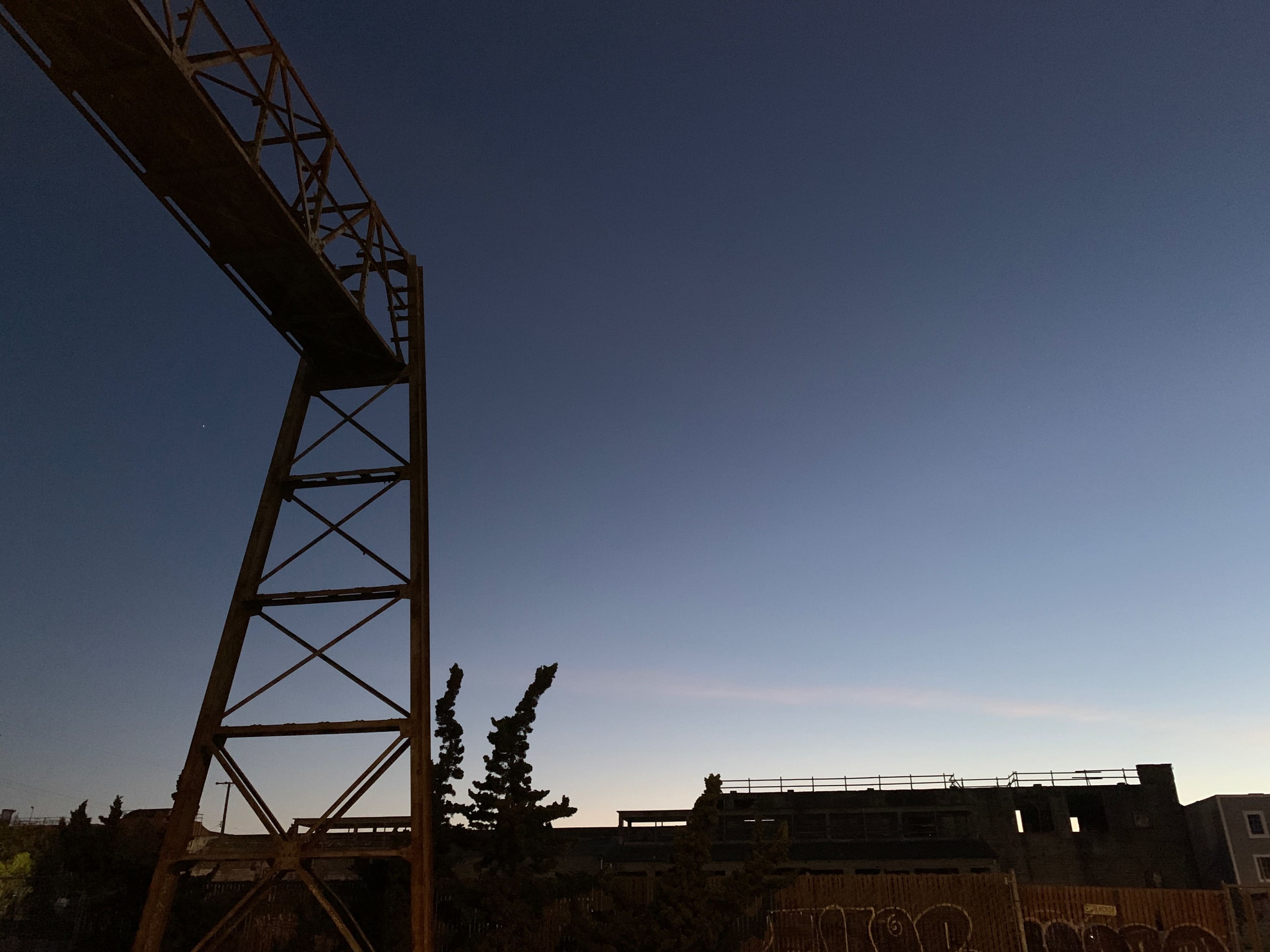 The image shows an industrial structure with a tall, rusted framework against a twilight sky, accompanied by trees and remnants of buildings in the background.