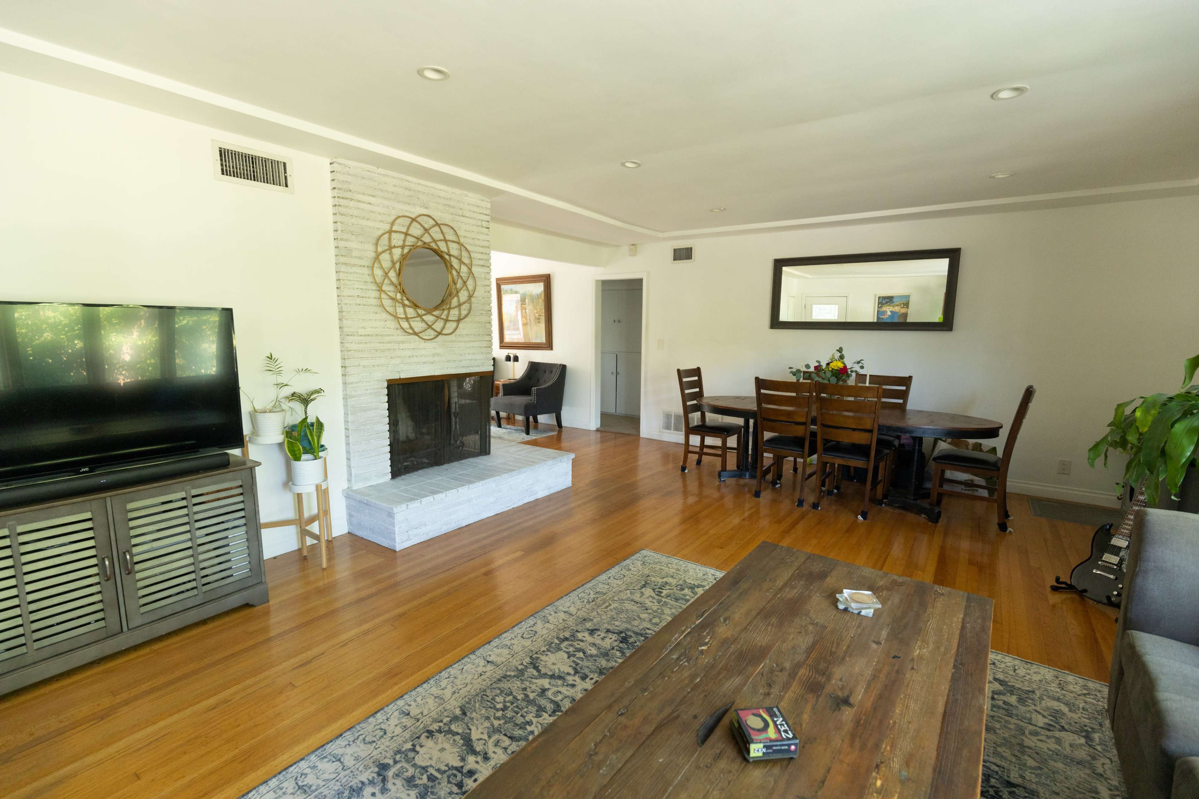 A living room with a television on a cabinet, a stone fireplace, a dining table with chairs, and a hardwood floor.