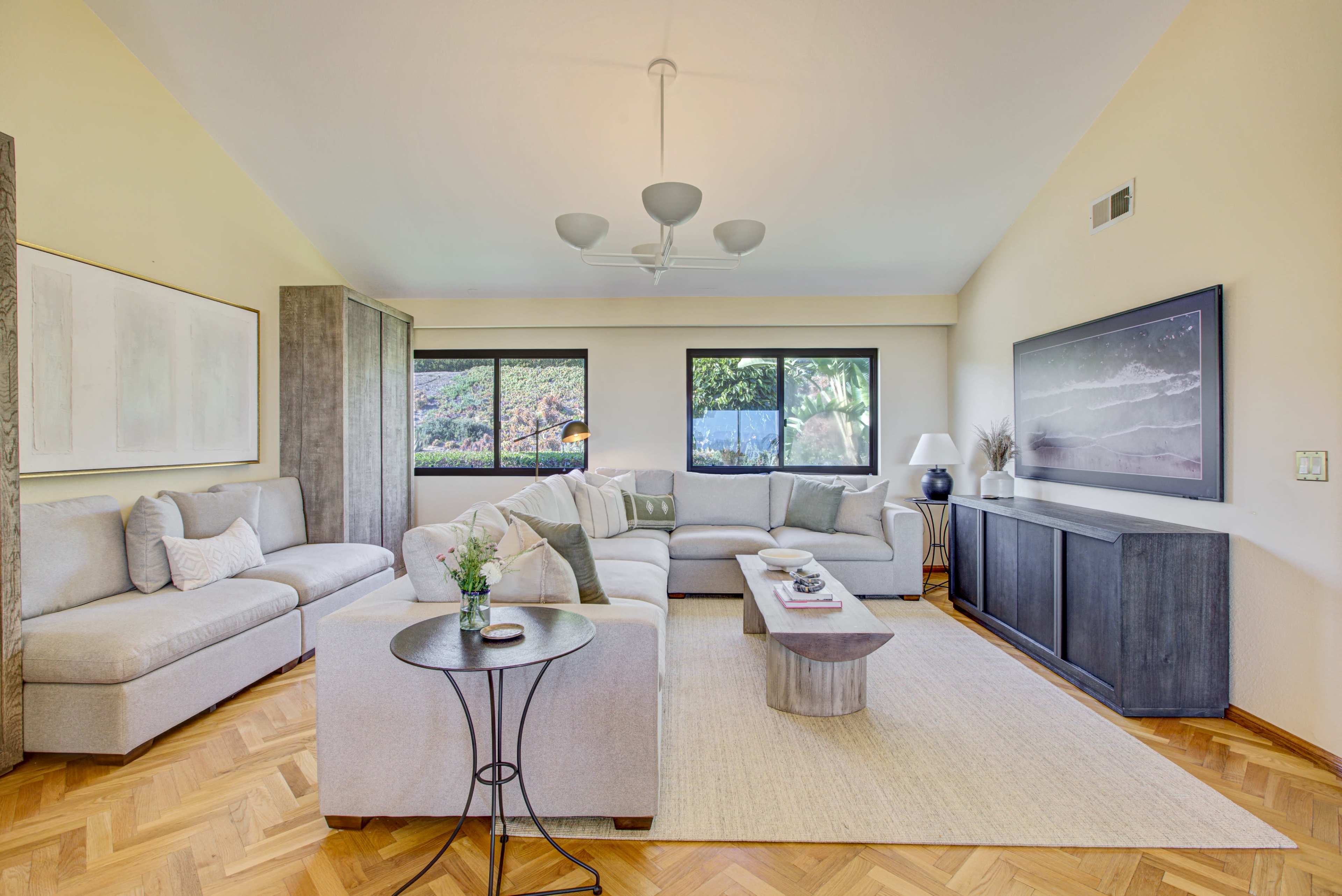 A modern living room with a light-colored sectional sofa, a wooden coffee table, and large windows that provide natural light.