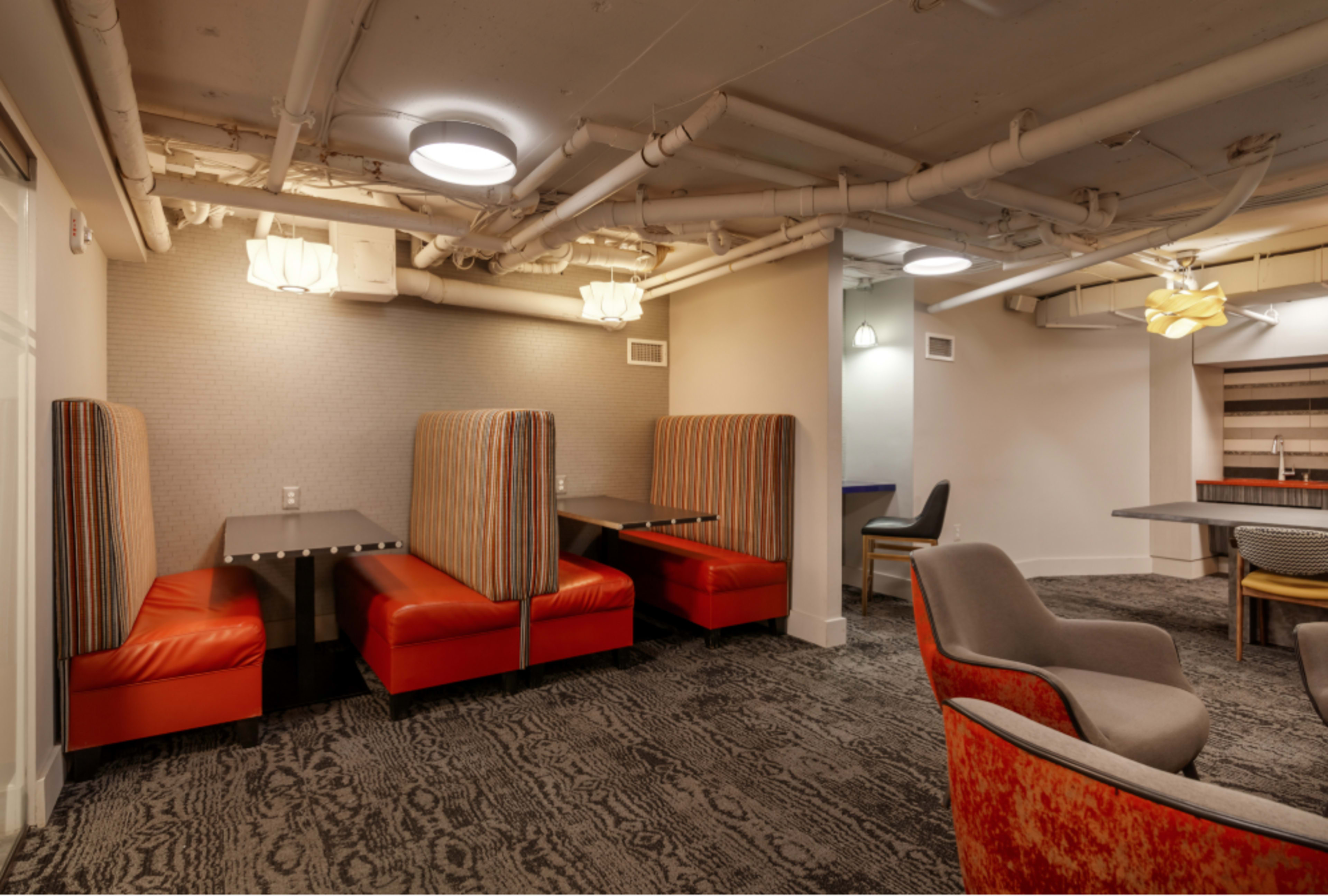 The image shows a lounge area with several seating booths and chairs, featuring striped upholstery and a mixture of table surfaces, all set beneath exposed piping and modern light fixtures.