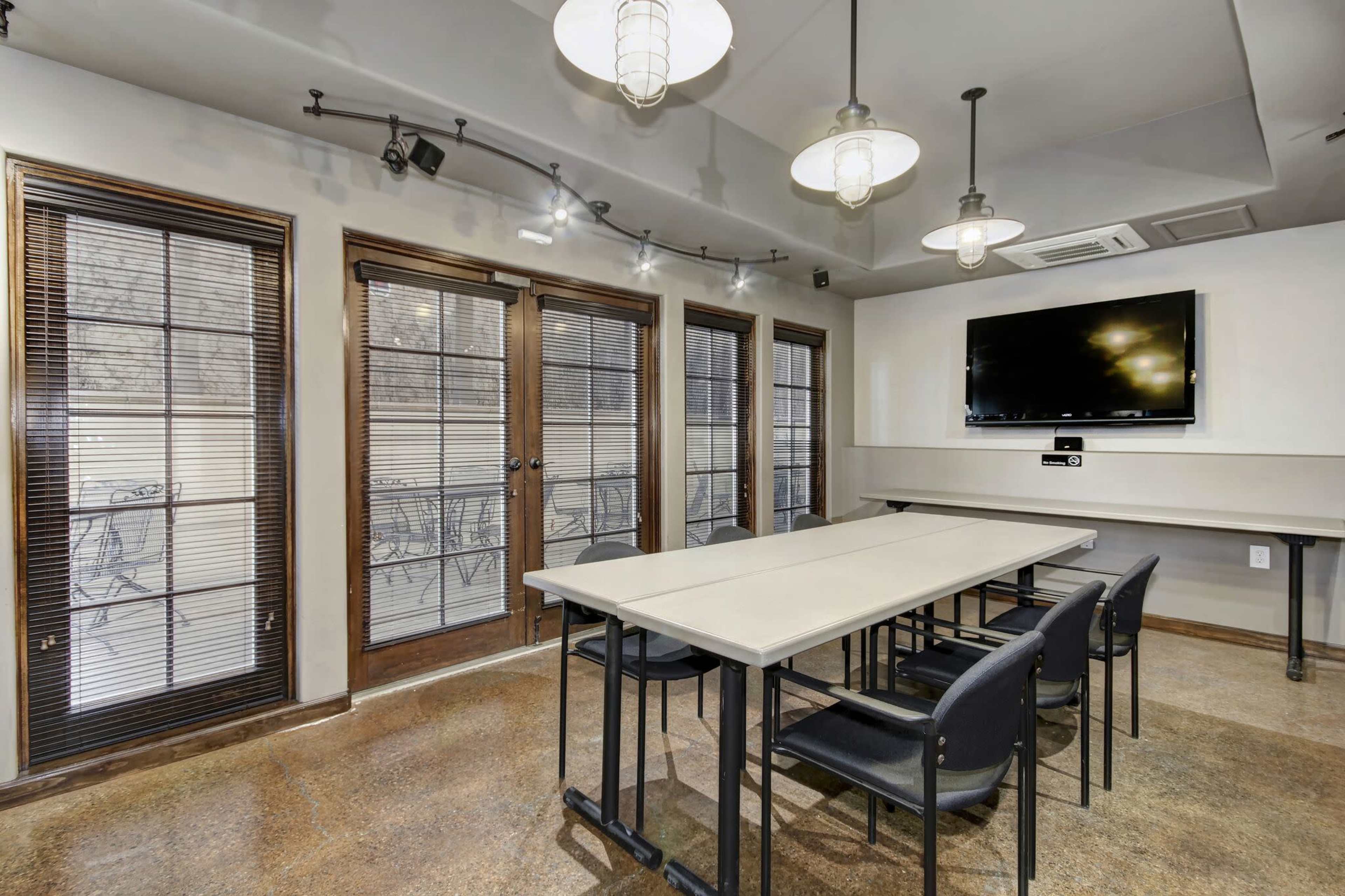 A meeting room features multiple tables and chairs, large windows with blinds, and a wall-mounted television.