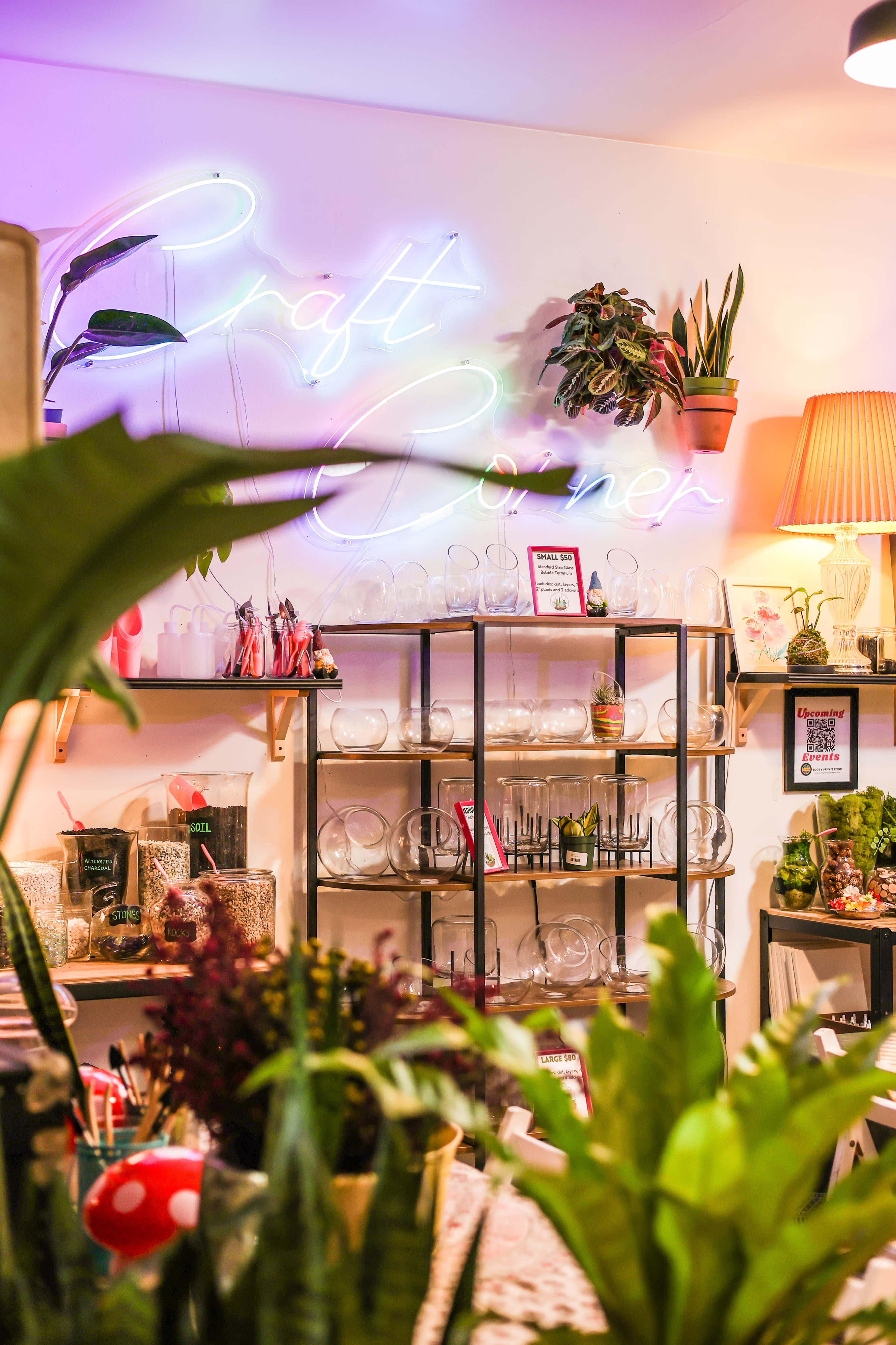 A colorful craft corner with a neon sign, shelves filled with glass containers, and various plants in decorative pots.
