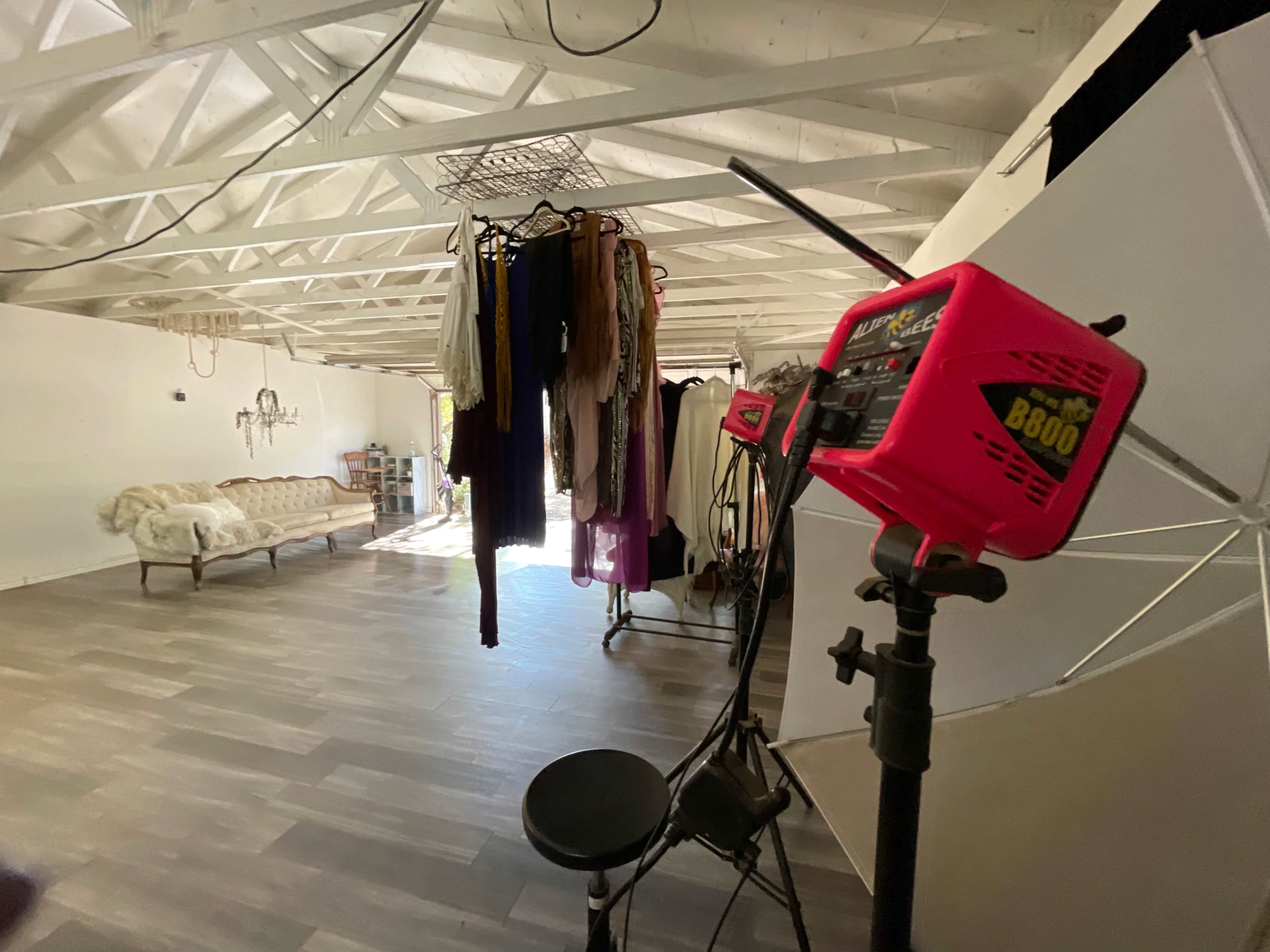 An open studio space with hanging clothing, a light stand, and a vintage couch against a white wall.