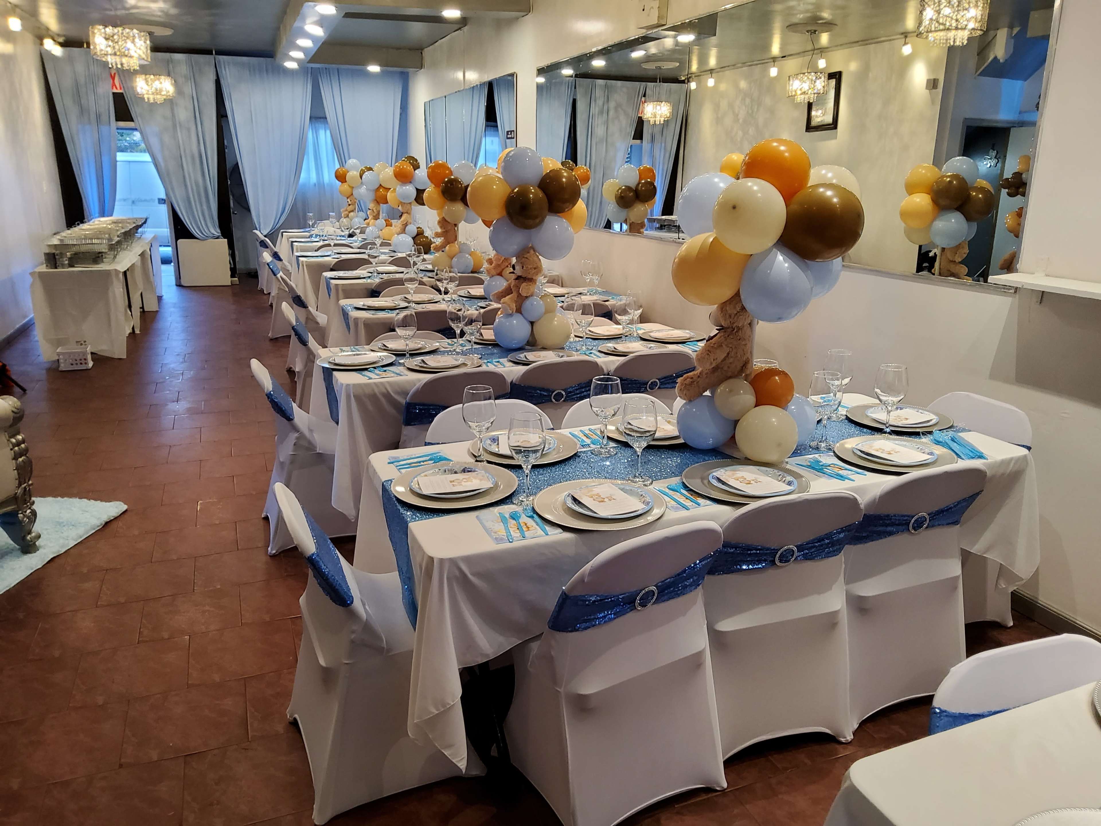 A decorated banquet hall with rectangular tables covered in white tablecloths, each set with plates, glasses, and blue decorations, flanked by balloon arrangements in various colors.