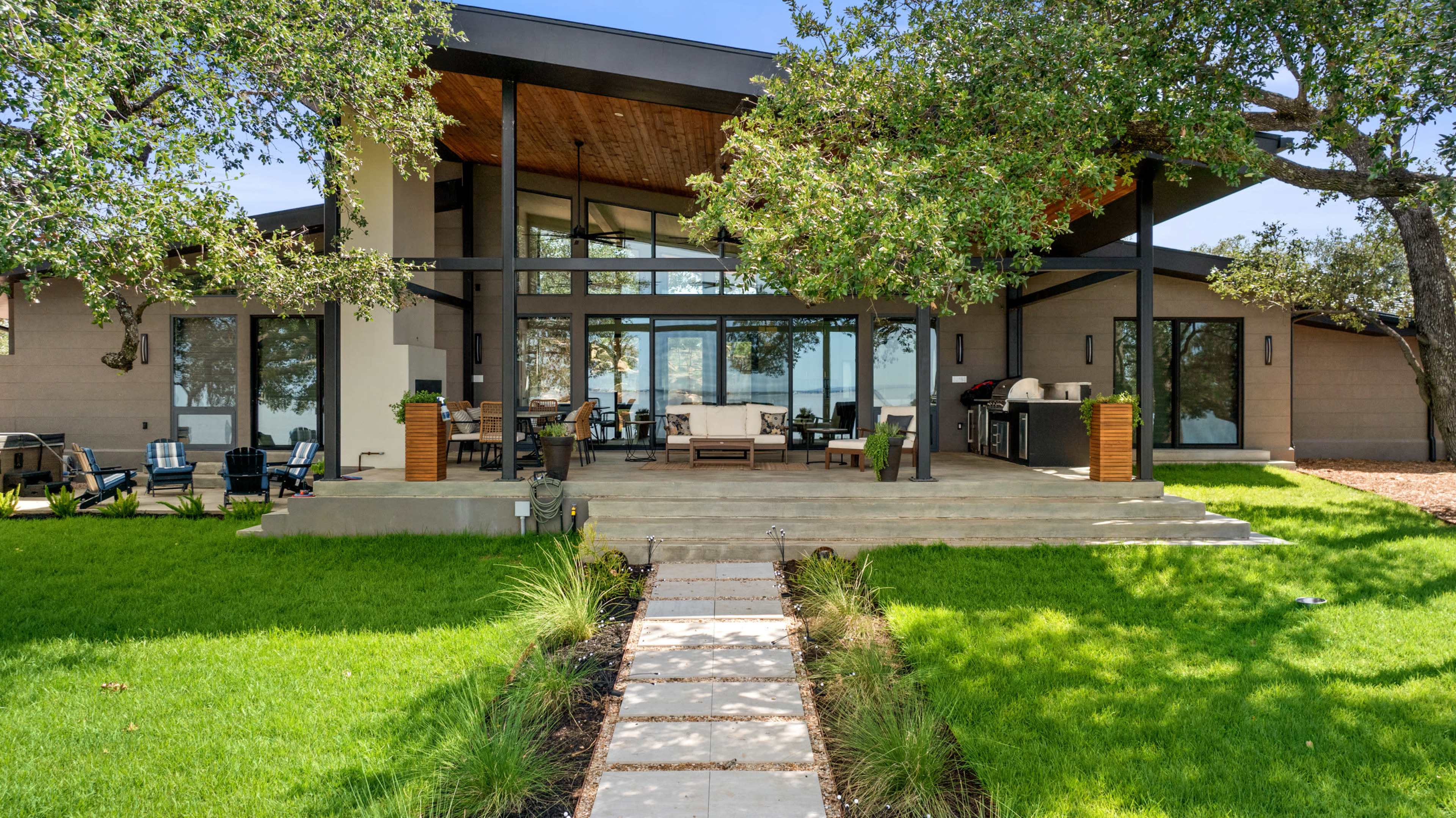 A modern house with large windows and a covered patio is situated on a grassy lawn, with a stone pathway leading to the entrance.
