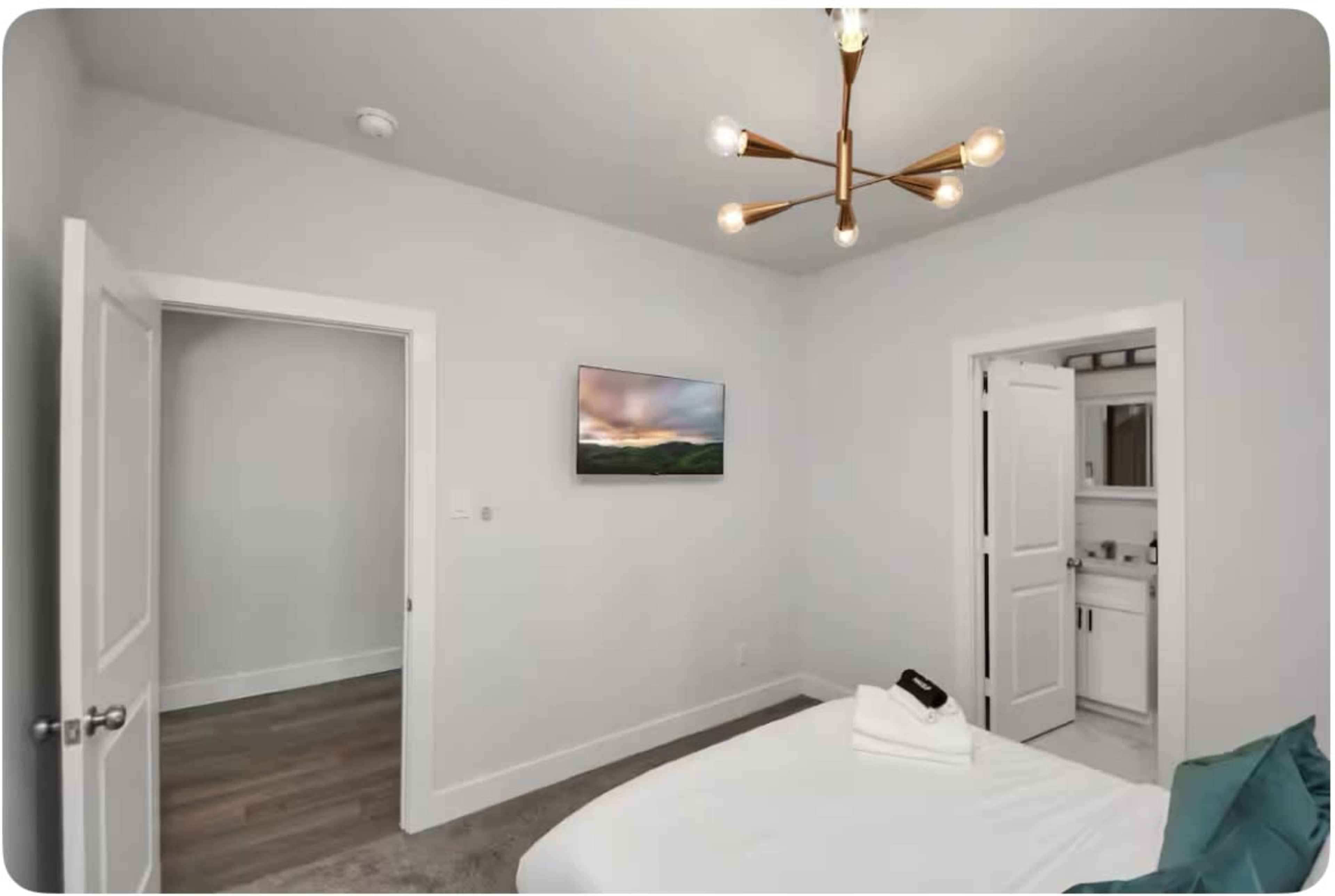 The image shows a minimalist bedroom with a bed, a wall-mounted television, and a door leading to a bathroom, illuminated by a modern light fixture.