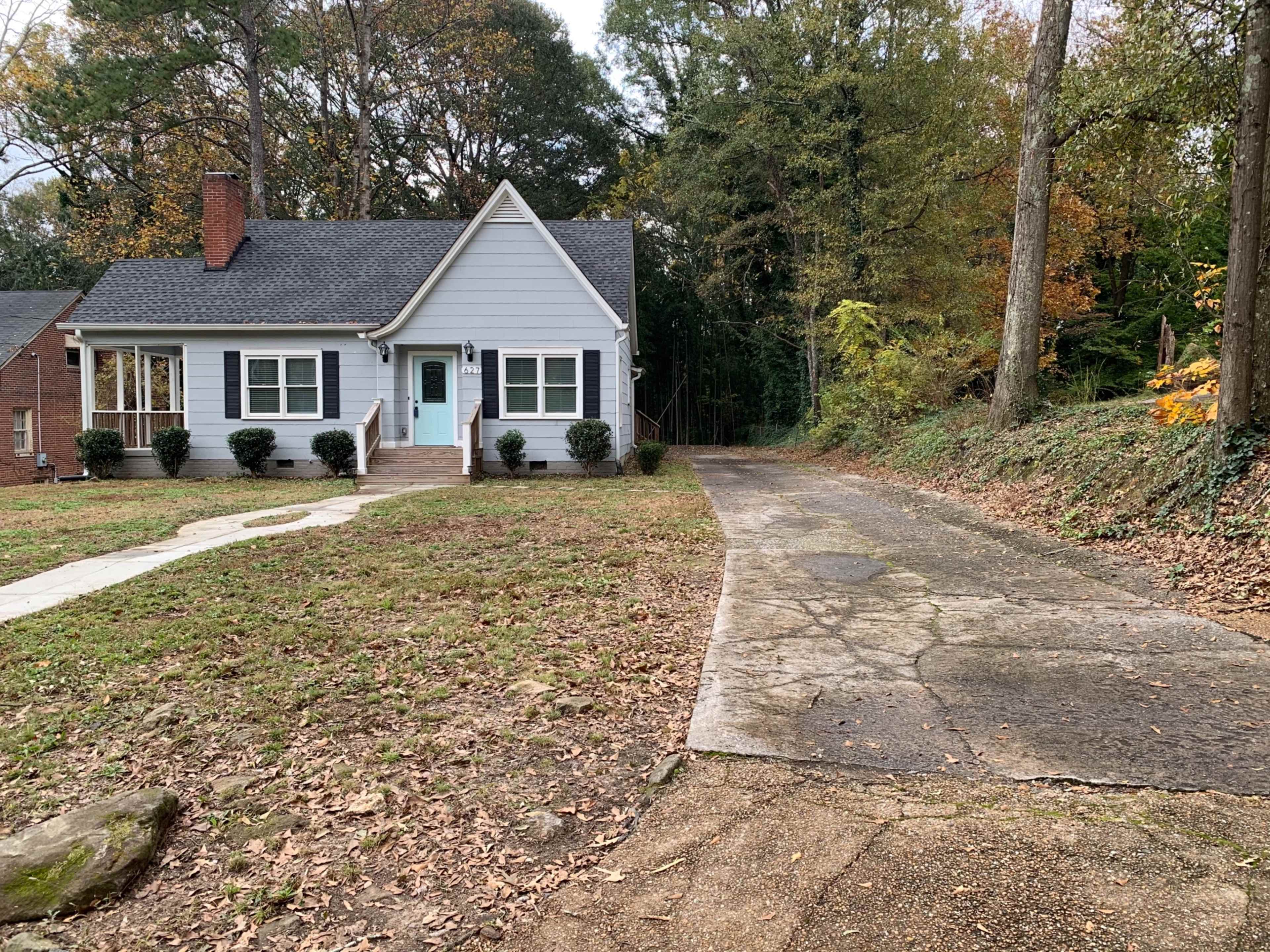A single-story blue house with a front porch, a neatly maintained lawn, and a gravel driveway alongside wooded foliage in the background.