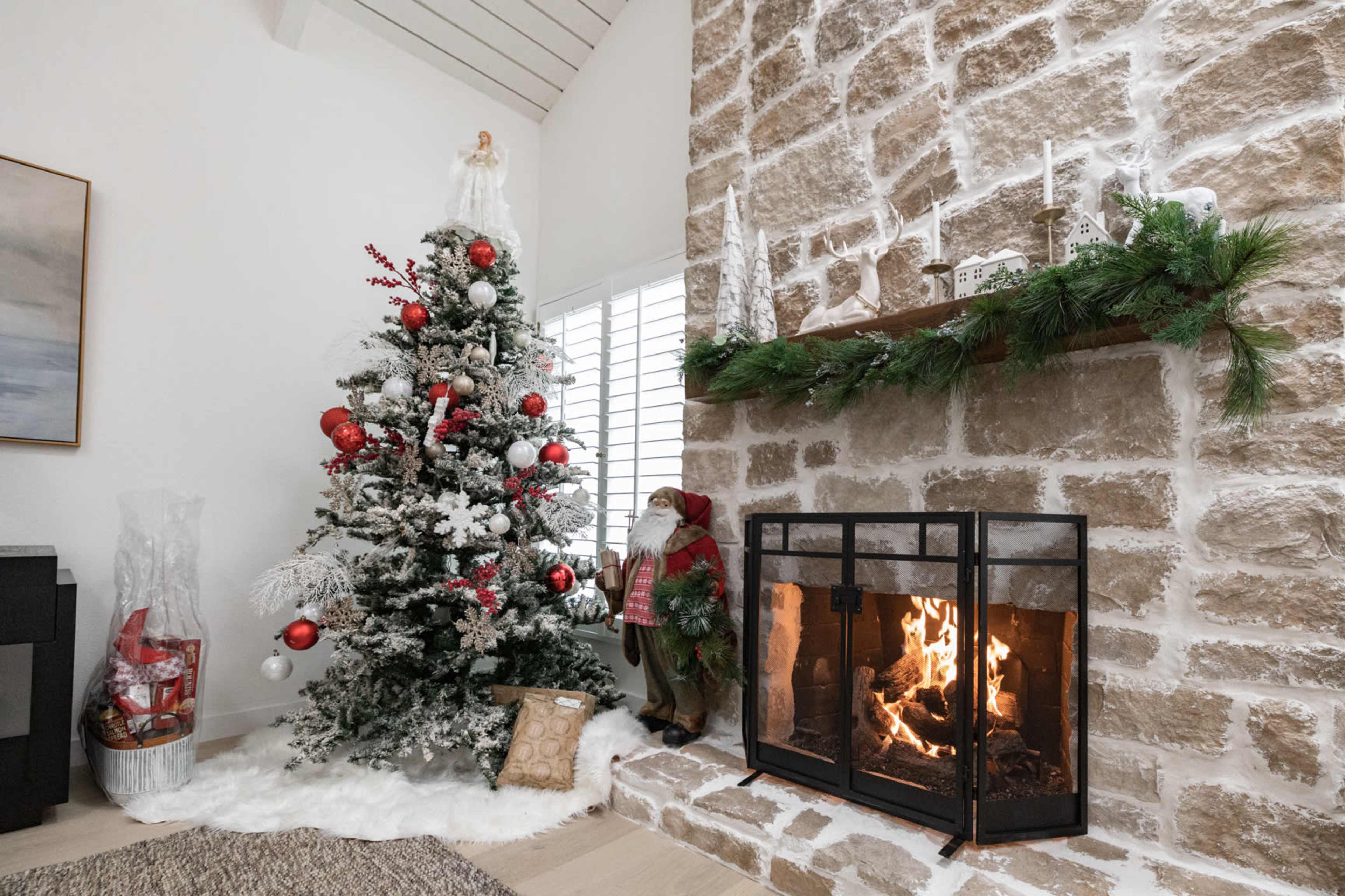 A decorated Christmas tree stands next to a stone fireplace with a fire burning, surrounded by holiday decorations and gifts.